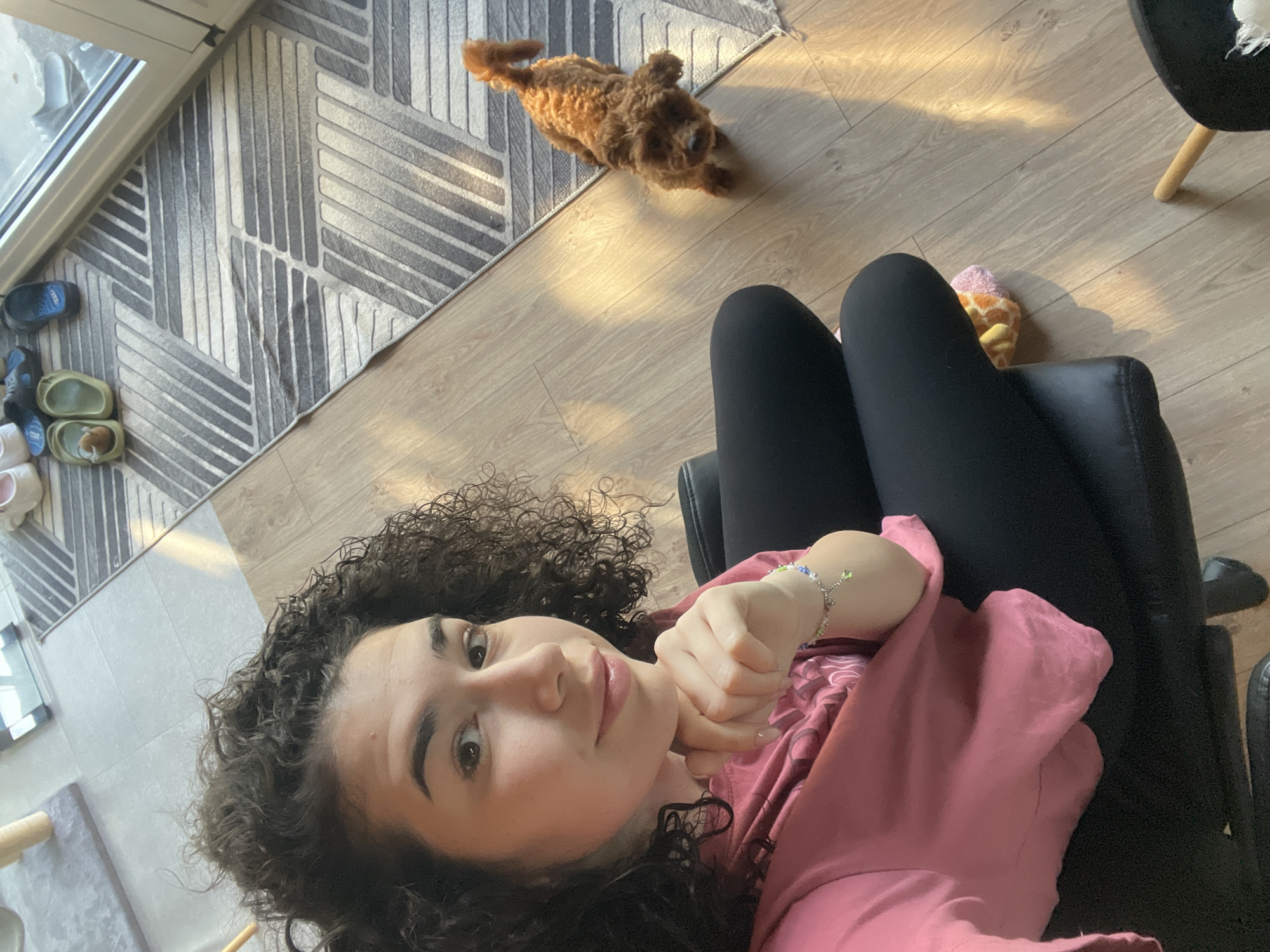 The image depicts a woman with curly dark hair, wearing a pink long-sleeved shirt and black leggings, sitting in a black chair. Her left hand is raised near her neck, showing a beaded bracelet. A brown poodle is lying on the wooden floor near a gray geometric patterned rug. There are shoes near the window, and a giraffe-patterned sock is visible near the chair. The specific location in Sofia, Bulgaria, cannot be determined from the given image.