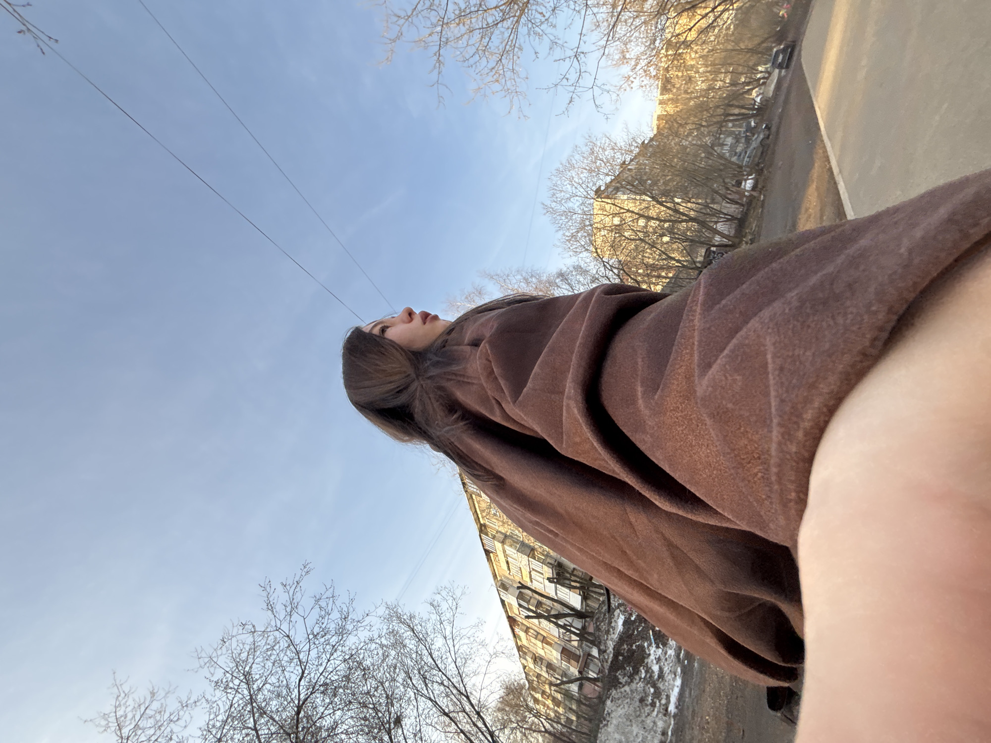 The image is a low-angle shot of a person outdoors, likely taken with a phone camera. The person, who appears to be a young woman with long brown hair, is wearing a brown coat or wrap. Her head is tilted upwards, her face visible in profile against a pale blue sky with some clouds. Above the woman, power lines and bare tree branches are visible against the sky.
In the background, buildings typical of an urban environment can be seen, likely residential or commercial. There are bare trees lining the street. A section of sidewalk and what seems to be part of a roadway are also visible. Based on the architectural style and the general appearance, it is likely this picture was taken in Perm, Russia. The presence of bare trees and patchy remnants of snow suggests it is either late winter or early spring. The light implies it is daytime. The person's arm and hand are in the foreground of the photo, suggesting a selfie or a photo taken by someone very close.
