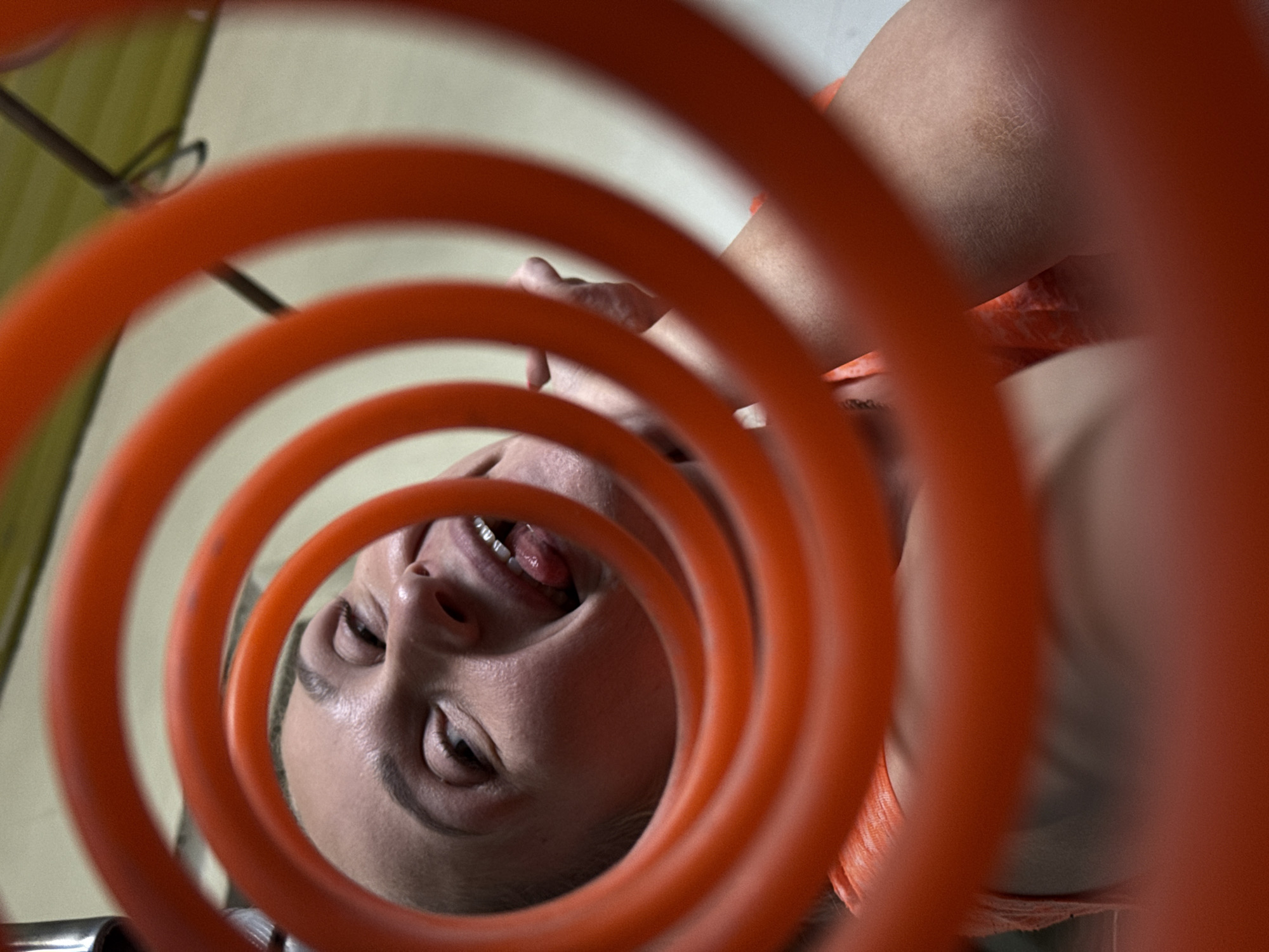 A person is viewed through an orange coiled tube. The person is lying down, their face is visible, and their tongue is sticking out slightly. The background is blurred, with hints of green and white surfaces. A metal rod is partially visible in the background. The location is not evident from the visual information provided, and there's no direct indication of it being Chaikovsky, Russia.