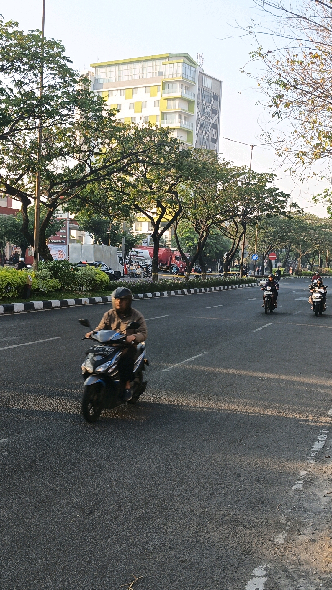 This is a vertical, eye-level, outdoor, daytime photograph taken from a street in Surabaya, Indonesia. In the foreground, a person wearing a helmet and a brown jacket is riding a blue motorcycle. The motorcycle has a license plate with blue lettering that is partially obscured. The rider is facing forward and appears to be looking ahead. The road is asphalt, and there are lane markings on the right side.
In the middle ground, several other motorcycles are also visible on the road, with riders wearing helmets. To the left of the road, there is a sidewalk with a black and white striped curb, followed by a median strip with manicured green bushes. Beyond the median, trees with green foliage line the street. In the background, a modern building with white and yellow accents and balconies stands tall. There are also other vehicles, including a red truck and a white truck, parked or moving in the distance. The sky is clear and bright.