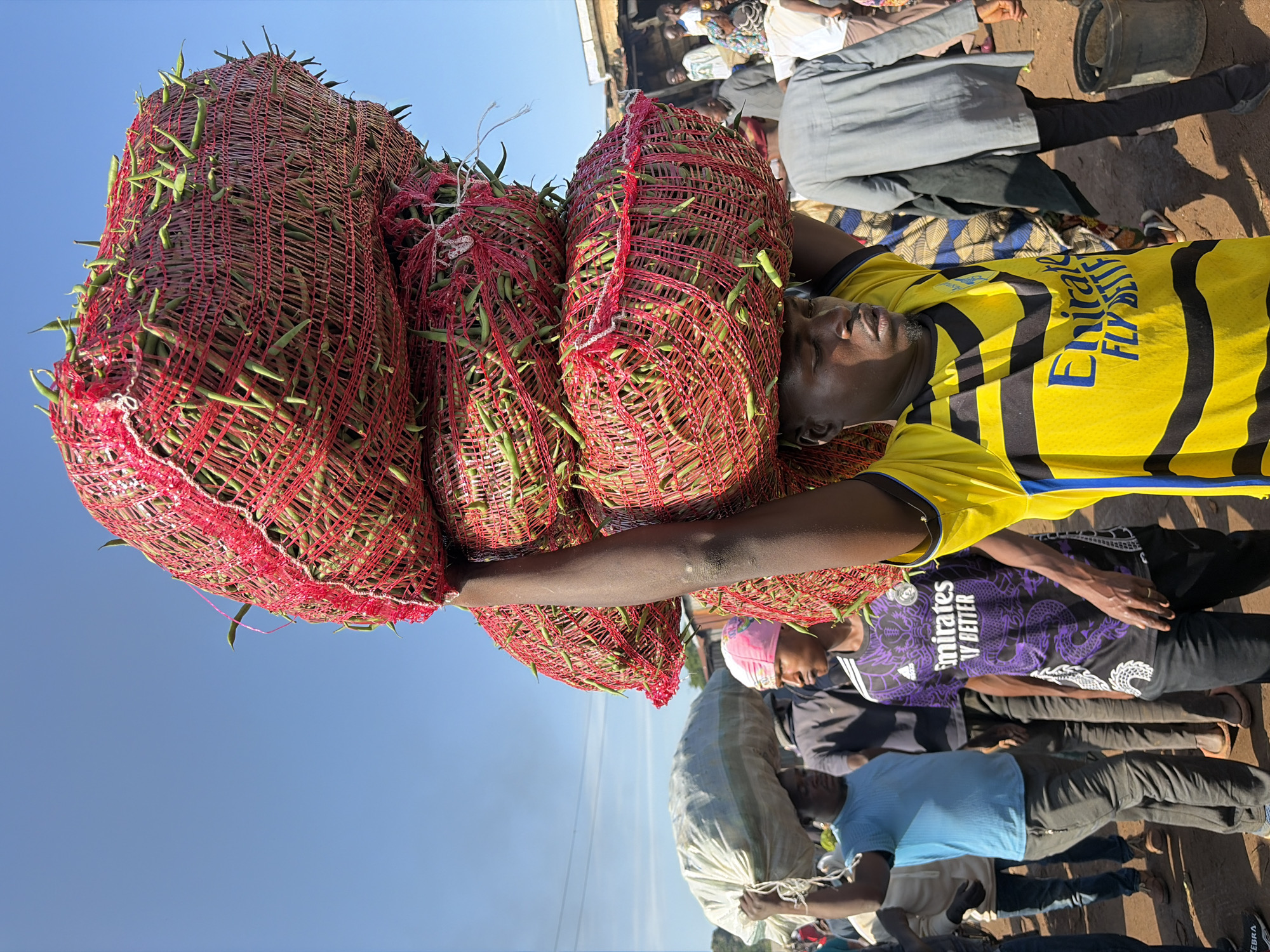 In Gura Top, Nigeria, a man carries a large stack of netted bags filled with green beans on his head. The bags are red and appear to be made of a woven material, with the green beans protruding from the mesh. He is wearing a yellow and black striped t-shirt. The scene takes place outdoors, likely in a market, with other people and stalls visible in the background. The sky is clear and blue.
