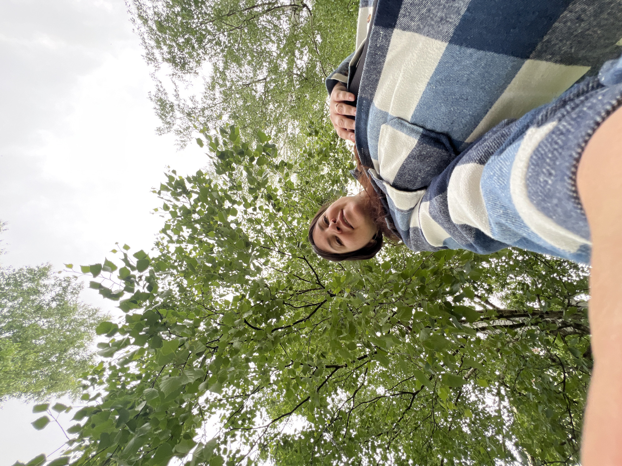 A low-angle shot, likely a selfie, features a woman wearing a blue and white plaid shirt. Overhead, the sky is visible through dense tree foliage. The location is Noviy, Russia.