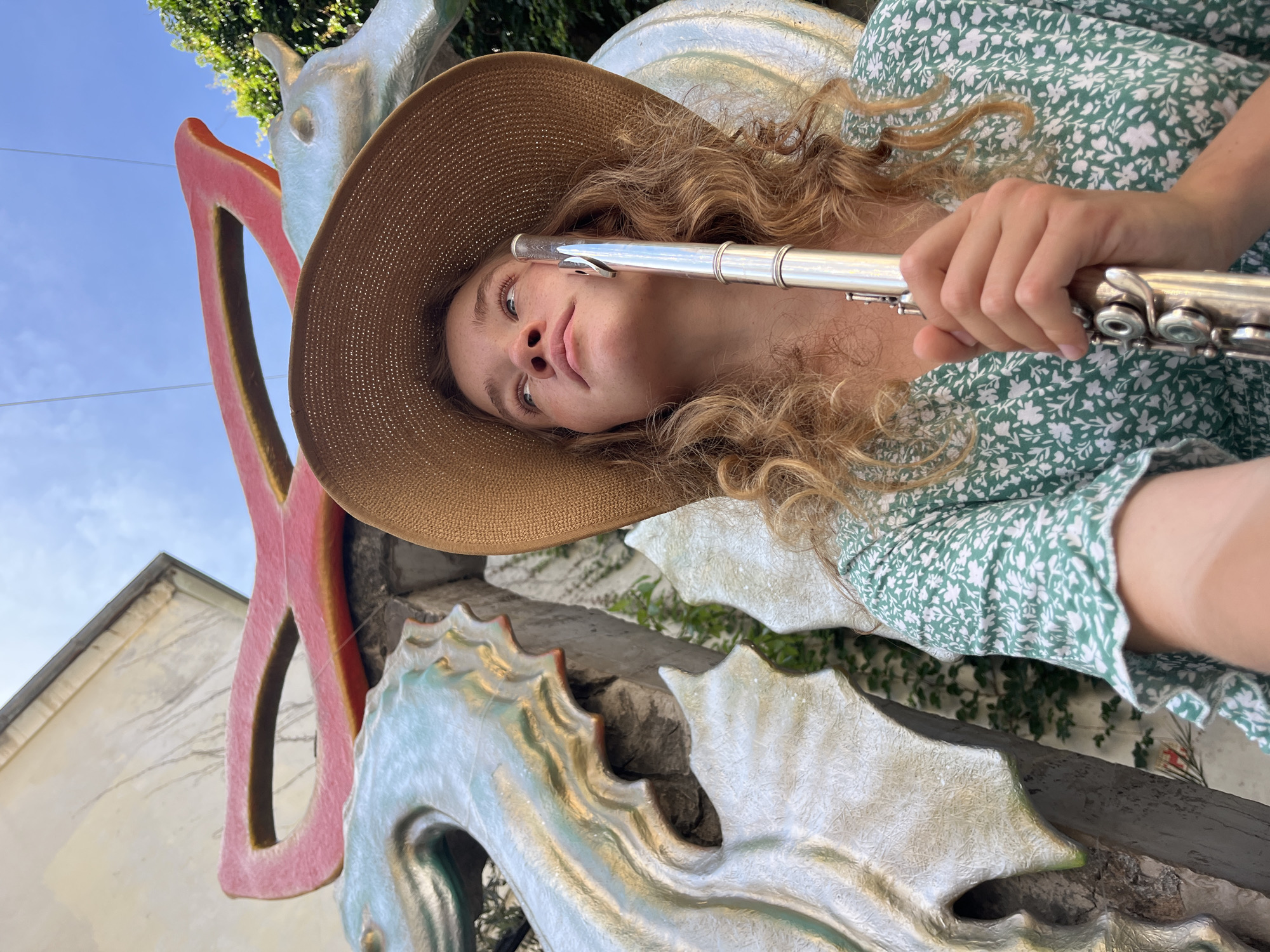 A young woman with long, wavy blonde hair, wearing a brown wide-brimmed straw hat and a green dress with a white floral pattern, is captured in a bright, rotated outdoor shot. She holds a silver flute near her mouth, looking upwards with a contemplative and calm expression.
The setting is an artistic outdoor space in Budva, Montenegro. Behind her, a large, light-colored sculpture or decorative architectural element is visible, featuring textured, wave-like or scale-like patterns with hints of white, light blue, and orange/red. A prominent, elongated, abstract red-painted structure stands behind her head, contrasting with the clear, bright blue sky and sparse clouds. Lush green foliage and parts of a light-colored building are also present in the background. The image is taken in daylight, suggesting a sunny day. The diagonal orientation of the photo creates a dynamic perspective. No text is visible.