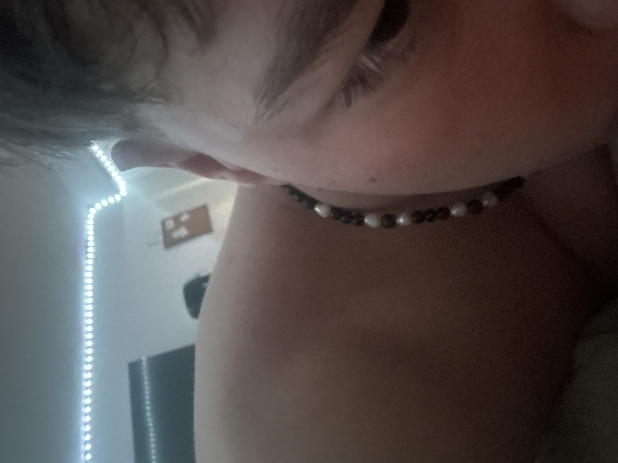The image displays a close-up, low-angle view of a person in an indoor setting. The subject, whose gender is not definitively identifiable, occupies the foreground, with the right side of their face, including the ear, eyebrow, and a partially obscured eye, visible. Their head appears tilted or they are in a reclining position. A beaded necklace or chain, composed of alternating dark and light-colored beads (resembling pearls), is worn around their neck/collarbone area. The skin tone is fair.
The background shows a light-colored room with a white ceiling. A strip of illuminated cool-white LED lights runs along what appears to be the perimeter of the ceiling. On a wall in the upper left background, a dark, rectangular object (possibly an electronic screen or television) is mounted. Further to the right, a lighter, rectangular object, possibly a framed item or decor, is also mounted on the wall. An indistinct dark shape is present in the mid-background, potentially another individual or furniture. The lighting is primarily artificial from the LED strip.
The specific location within the city of Troyan, Bulgaria, cannot be confirmed from the visual information presented in the image, as the environment lacks distinctive geographical or architectural identifiers. The scene depicts a generic interior.