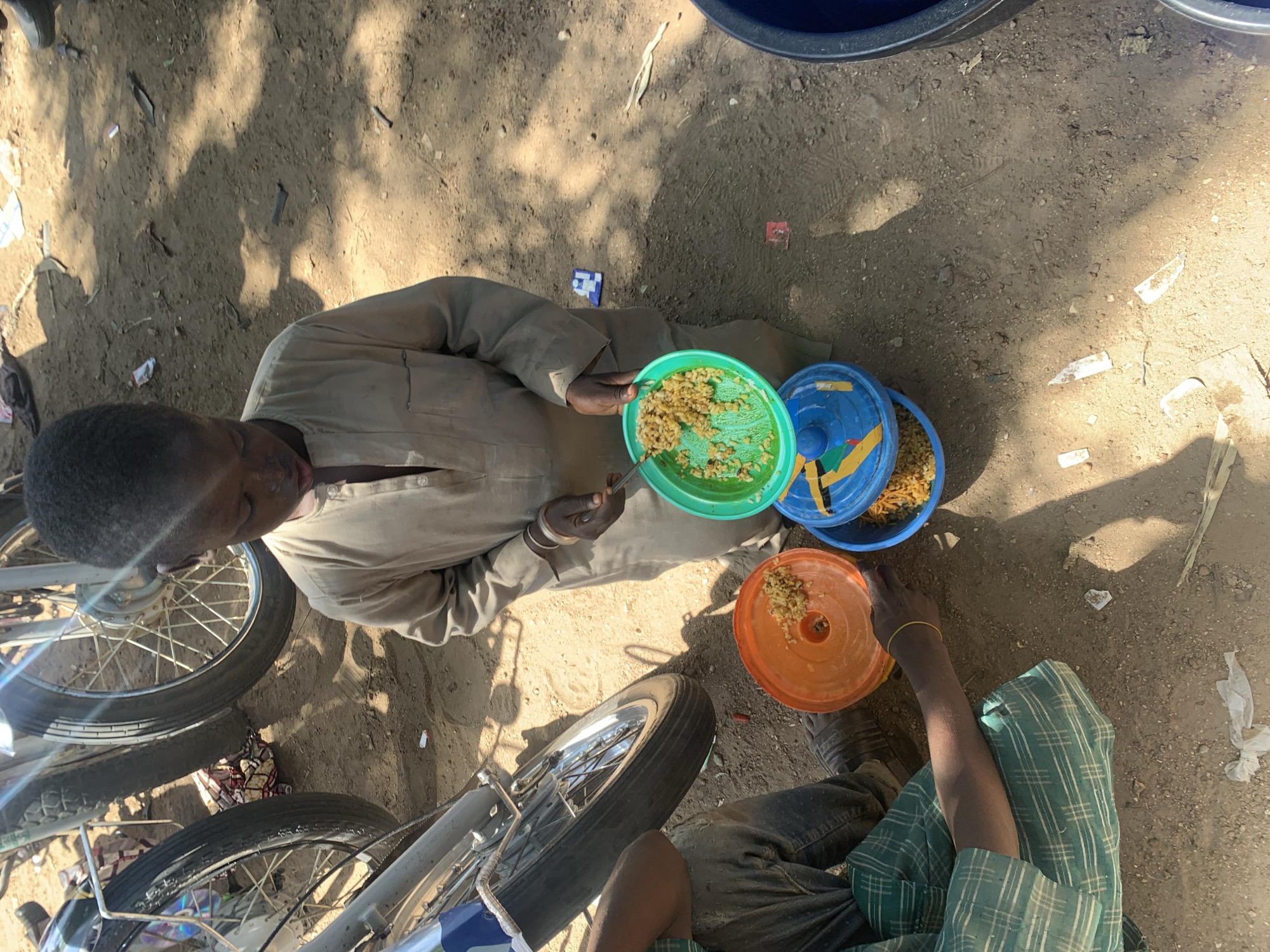A young boy in Boi A, Nigeria, holds a green plate filled with food. He is wearing a tan tunic. In front of him are three plastic containers, one orange, one blue, and another one with a blue lid. The orange container has food in it. The boy appears to be eating. He is standing on a dirt ground. Two motorcycles are parked behind him. There is also a person sitting next to the boy wearing a green and white checkered shirt.