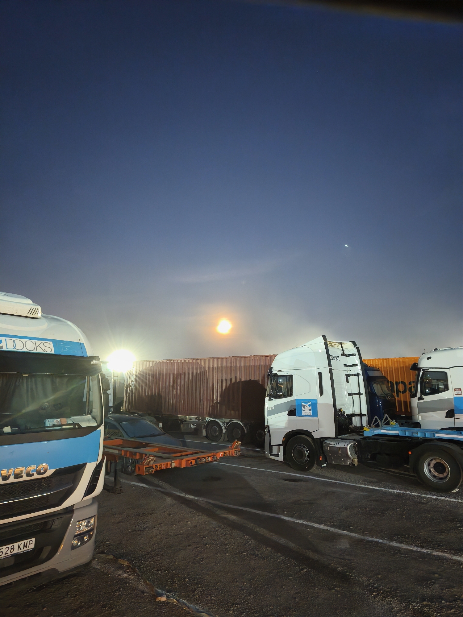 The image shows a truck stop at night in Alcàsser, Spain. Three large white trucks, two of which have "DOCKS" written on their sides, are parked on an asphalt surface marked with white lines. One truck is pulling a flatbed trailer, upon which sits a dark-colored car. Behind the trucks are shipping containers. The sky is dark blue with a bright moon or streetlight visible in the distance. The scene is illuminated by artificial lights, creating long shadows.