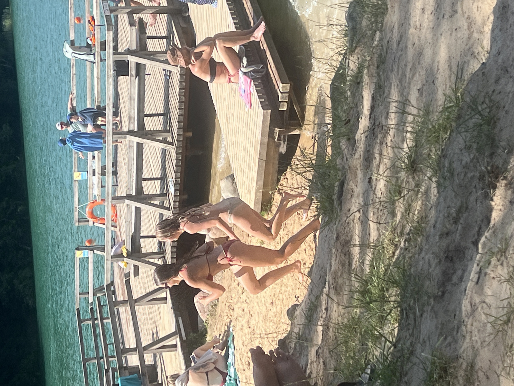 A group of young women are enjoying a summer day at a lake in Vilnius, Lithuania. They are dressed in bikinis and appear to be sunbathing and socializing on a wooden pier and the sandy shore. The water is a vibrant turquoise, suggesting it is a clear day with good weather. The sun is high in the sky, indicating it is likely midday or early afternoon.
On the pier, one woman is sitting with her legs up, looking to her left. Another woman is leaning forward, seemingly in conversation with someone on the pier. Further down the pier, two other women are in a more active pose, one appearing to be reaching out or jumping into the water. There are several life preservers visible on the pier, along with an orange ball.
The scene is set on a lake with a wooden structure that serves as a pier or dock. The shoreline is sandy with sparse vegetation. The overall mood seems relaxed and joyful, typical of a summer outing. There is no visible text in the image.