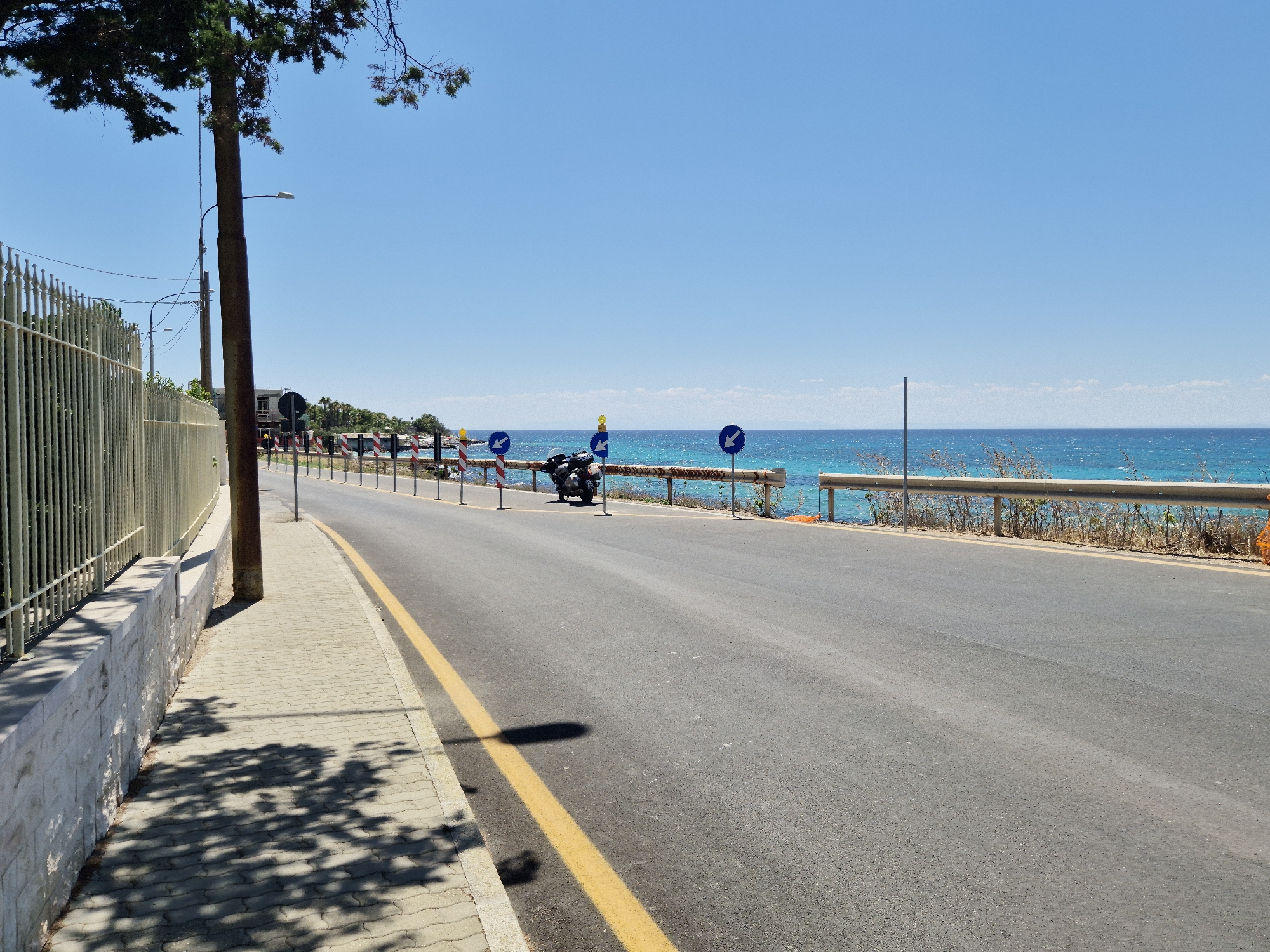 This is a roadside scene in Taranto, Italy, featuring a paved road running alongside a sidewalk and a metal fence. The road has a yellow dividing line. A dark-colored motorcycle is parked on the side of the road, near several blue directional signs. The sea is visible in the background, with clear blue water under a bright sky. Trees and vegetation are also present. The scene appears to be a sunny day.