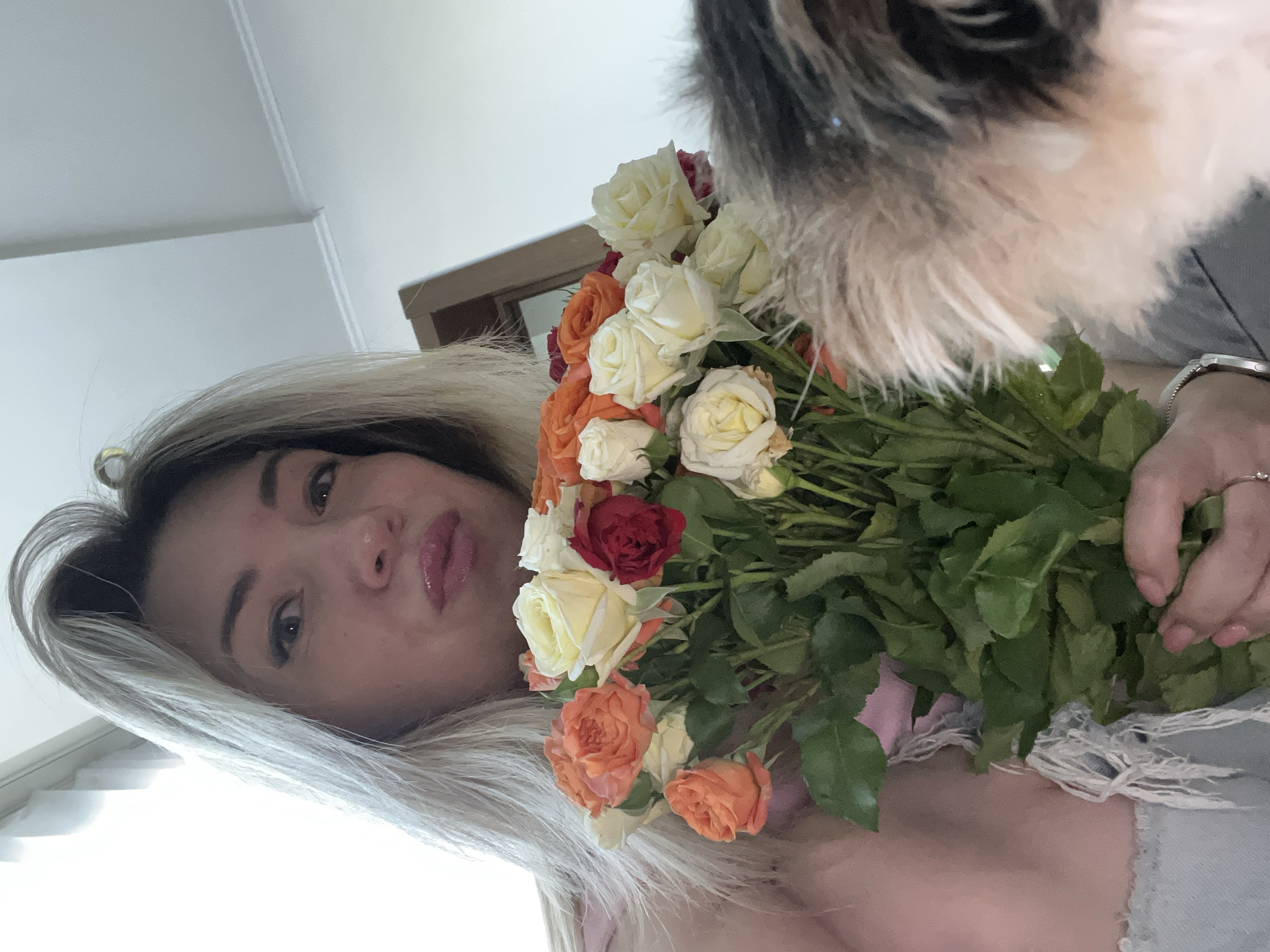 A selfie-style, close-up shot features a woman with blonde hair holding a bouquet of roses. The woman's face is partially obscured by the flowers, but her lips are slightly puckered. She is wearing a pink top and a silver watch. To the right of the woman, a fluffy animal, likely a cat, is partially visible. The background appears to be an indoor setting with white walls and a ceiling. The lighting suggests it is daytime, with natural light coming from a window. The scene does not contain any elements that place it specifically in Sochi, Russia, or any other identifiable city.