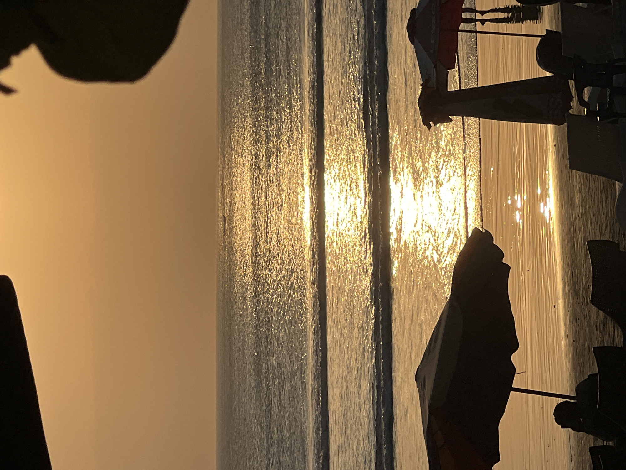 This is a photograph of Kuta Beach in Kuta, Indonesia, at sunset. The sun is low in the sky, casting a golden light across the water and the sand. Gentle waves are rolling in from the ocean, and the water is reflecting the warm hues of the sunset. In the foreground, there are silhouettes of beach umbrellas and lounge chairs, suggesting a relaxed beach scene. A few people are visible in the distance, wading in the shallow water or walking along the shoreline, also in silhouette. The overall atmosphere is tranquil and serene, with the natural beauty of the beach illuminated by the setting sun.