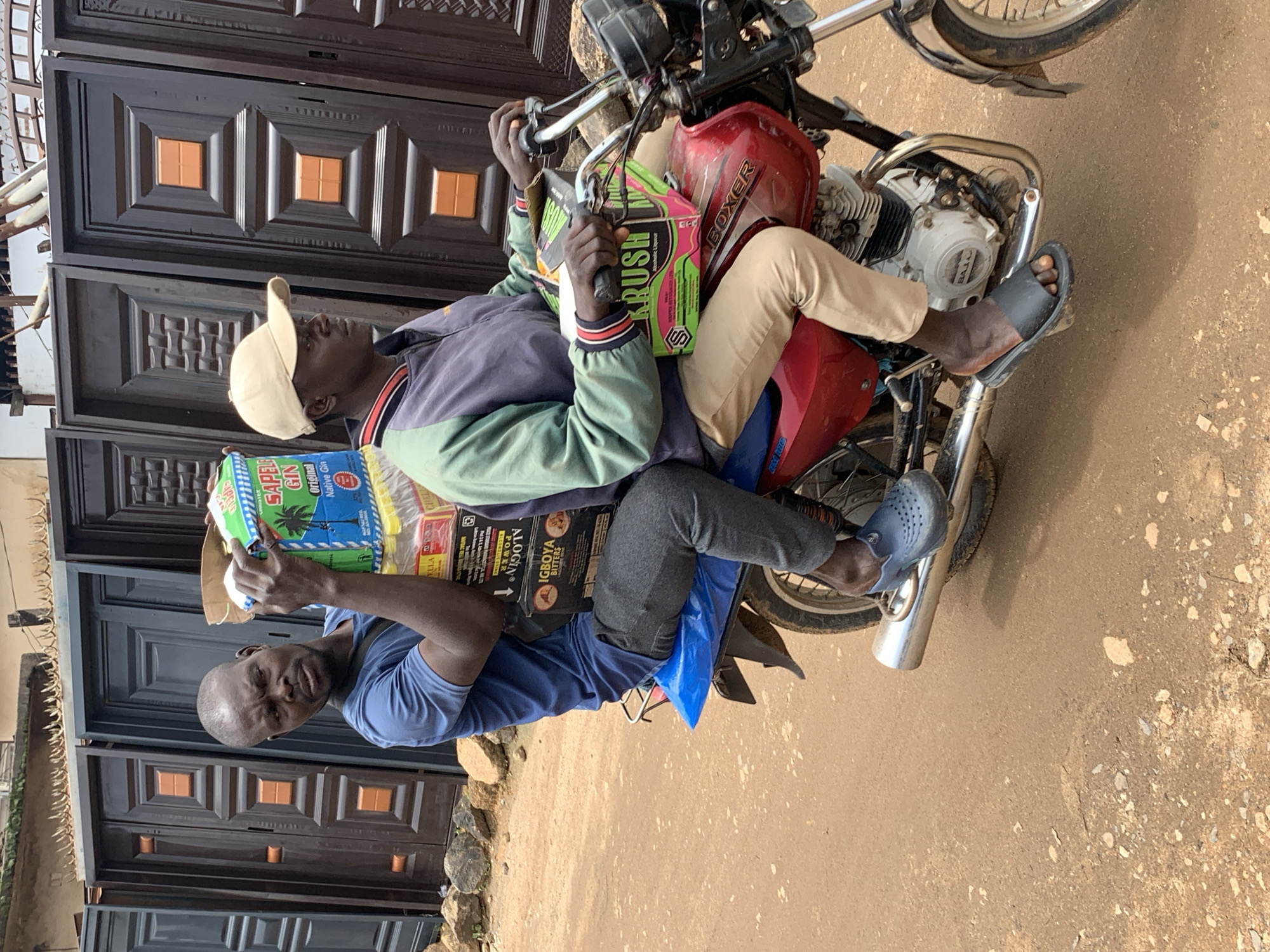 Two men are on a red motorcycle, with the driver wearing a light-colored baseball cap, a multi-colored jacket, and khaki pants. The man behind him, who is holding a stack of boxes and bottles, wears a blue t-shirt and dark pants. The boxes are green and yellow, with "SAPELE GIN" visible on one. The motorcycle is parked on a dirt road in front of a row of dark-colored metal doors. The background shows a building with a corrugated metal roof and what appears to be wire fencing. The scene is consistent with a market or roadside stall area in Gura Top, Nigeria.