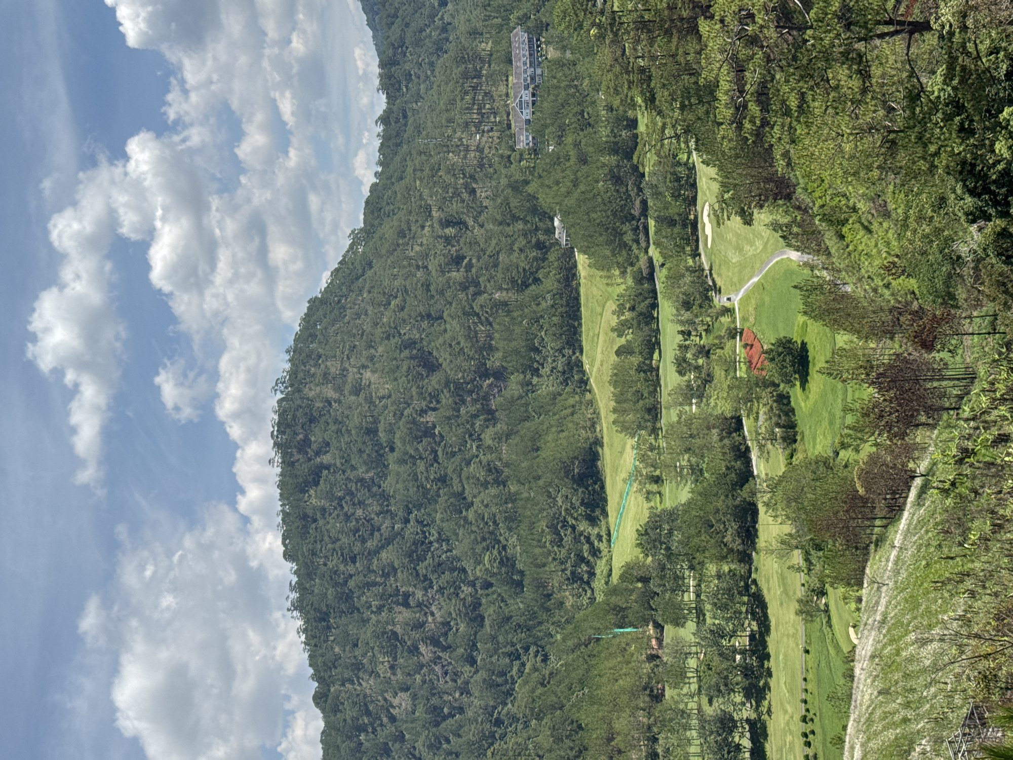 The image captures a landscape in Xuan Huong, Da Lat, Vietnam, featuring a hillside covered in dense green trees. A golf course with manicured green fairways and sand traps meanders through the trees. A few buildings, including what appears to be a large hotel or resort, are visible atop the hillside. The sky is light blue and filled with fluffy white clouds.