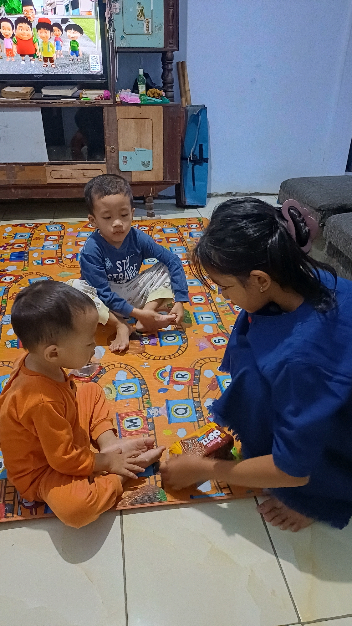 In a living room in Kowangan, Indonesia, three young children are gathered on a colorful play mat. The youngest child, wearing an orange outfit, sits closest to the camera, facing left. A slightly older boy in a blue long-sleeved shirt and patterned pants is seated behind the youngest, facing forward. An older girl, also in a blue outfit, is kneeling on the right, facing the younger children. She holds a snack package, and the children appear to be interacting around it, possibly sharing or discussing the food. The background reveals a television displaying a cartoon, a wooden cabinet, and a blue bag. The floor is tiled. The scene depicts a typical domestic moment of children playing and snacking in their home.