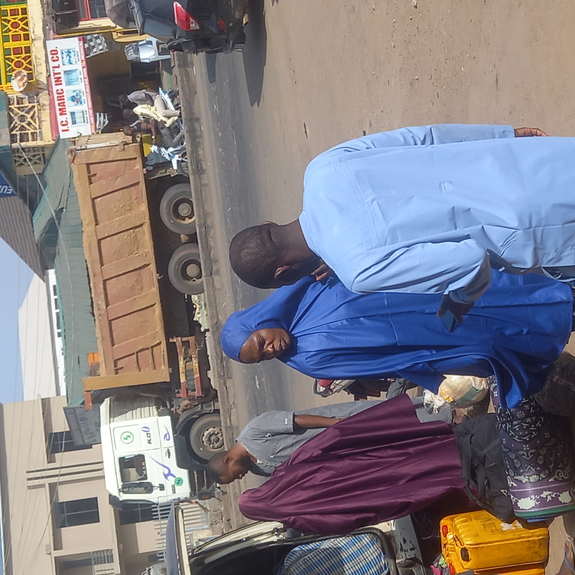 The image depicts a vibrant street scene in what appears to be an urban or commercial area, consistent with Sardauna, Nigeria, captured during a bright, dry day. The image shows a moment of everyday activity, possibly involving travel or waiting for transport.
In the foreground, several people are visible. One individual, seen from behind, wears a light blue traditional garment (likely a kaftan or bubu). Facing the camera, another person, appearing to be a woman, is dressed in a dark blue head covering and matching traditional attire, with a neutral expression. Adjacent to them are various items, including patterned bags, bundled textiles, and a yellow jerry can, suggesting they may be traveling or awaiting transportation. Slightly behind them, another person in a grey shirt is also present.
The background features a road with multiple vehicles and buildings. A large, rusty brown dump truck is prominent on the street, and further down, the white cab of a MAN truck, marked with "K00" and "MAN," is visible. A black car is partially seen towards the upper left.
On the left side of the street, buildings line the roadside. One building features a distinctive signboard for "I.C. MARC INT'L CO.," which displays images of household appliances like refrigerators and air conditioners, indicating it's an electronics or appliance store. Additional text on the sign mentions "WHOLESALE RETAILERS OF." The ground appears dusty, and the bright sunlight suggests it is mid-day or afternoon. The overall scene conveys a sense of daily life and commerce in an active town environment.