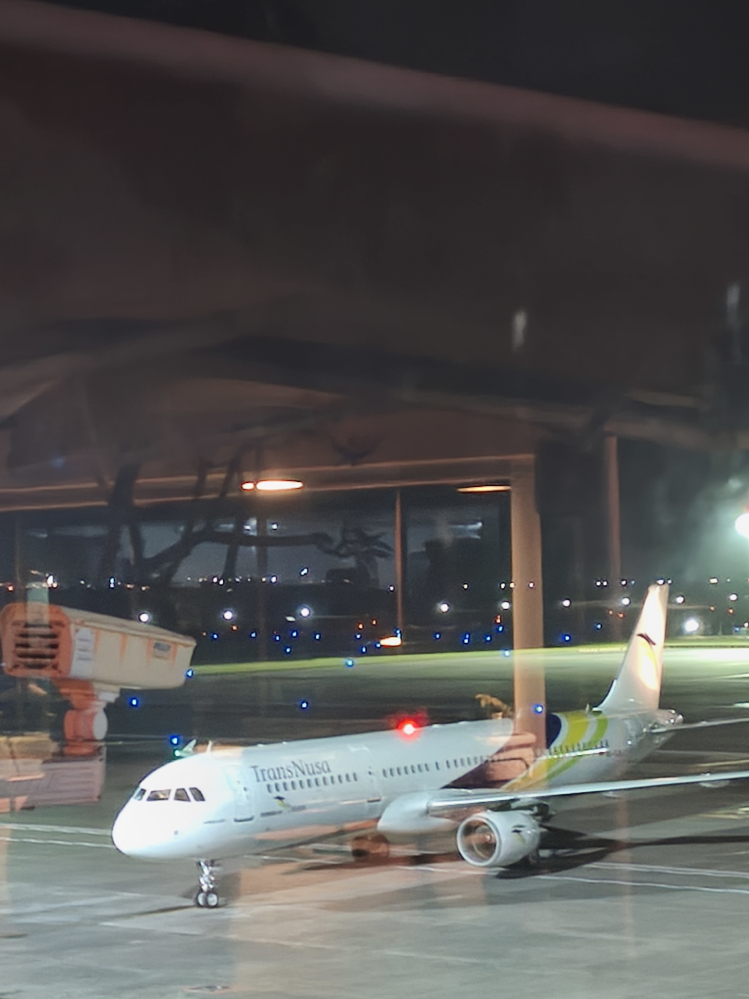 The image depicts a TransNusa airplane parked on the tarmac at an airport at night. The airplane is white with a colorful tail design featuring yellow, green, and blue. The cockpit windows are visible, as are the engines. The tarmac is illuminated by runway lights, which appear as blue and white dots. In the background, there are airport structures and lights that suggest the presence of a terminal building. To the left of the airplane, there is an orange and white ground support equipment. The scene is captured from a slightly elevated perspective, possibly from within the airport terminal, looking out through a window. The overall impression is that of an airport at night, with an airplane ready for departure or having just arrived. The location is identified as Kuta, Indonesia.