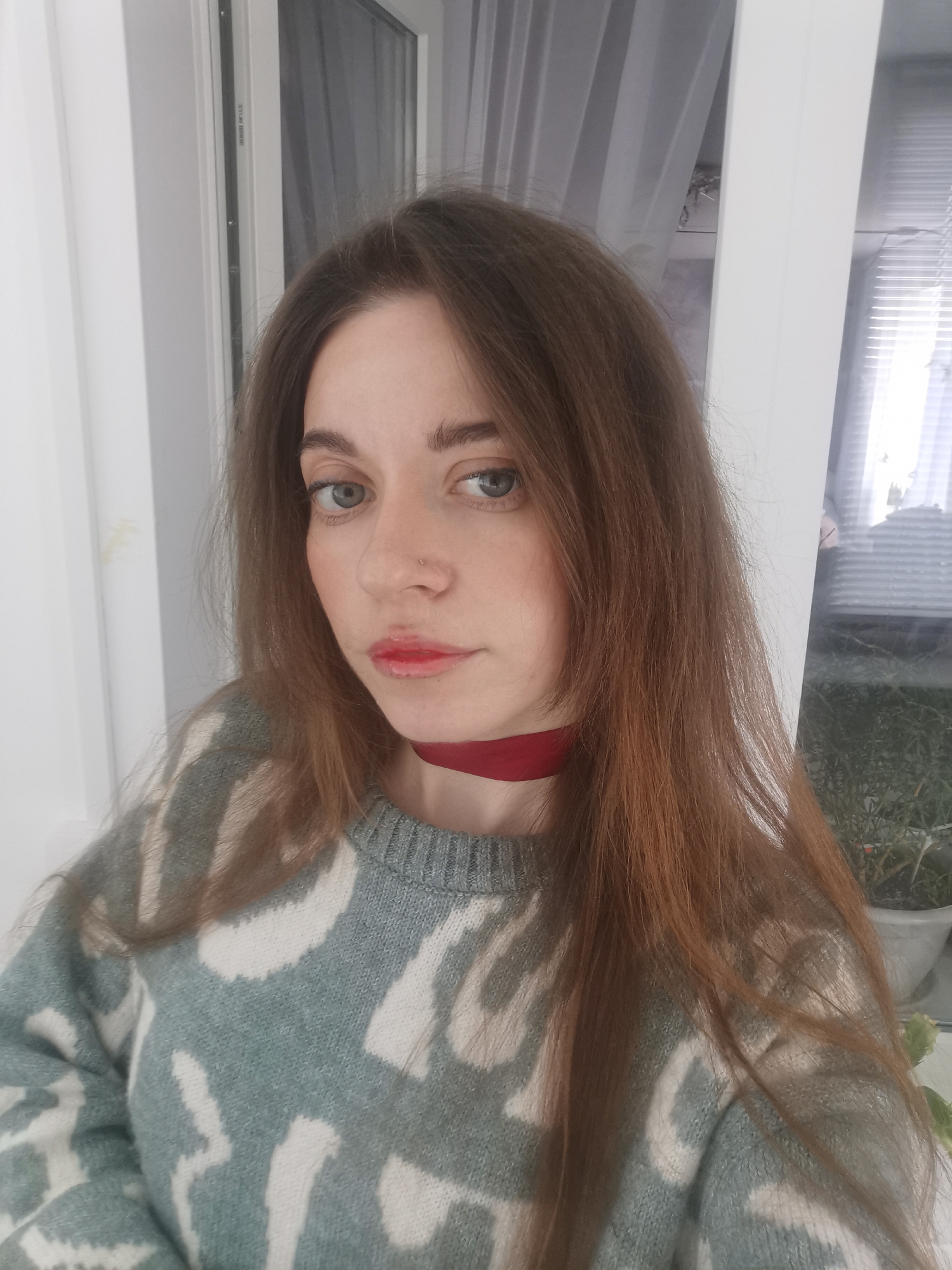 A young woman is depicted in a close-up, chest-up portrait, looking directly at the camera. She has long, straight, reddish-brown hair, parted in the middle, framing her face. Her eyes are light-colored, appearing blue or green, and she has a small nose piercing on her right nostril. Her lips are partially covered with red lipstick, which appears somewhat uneven or faded towards the center of her upper lip.
She is wearing a crew-neck knit sweater in a light blue or teal color with a white, abstract, or camouflage-like pattern. Around her neck, she wears a thin, dark red ribbon or choker. Her expression is neutral and calm.
The setting appears to be indoors, likely in front of a large window or glass door. The background on her left is a plain white vertical frame, while on her right, there's another white vertical frame, and behind it, partially visible sheer white or light gray curtains. Beyond the curtains, hints of blurred greenery, possibly potted plants or outdoor foliage in a container, are visible on the right side of the image, suggesting daytime.
Faint, dark text is visible vertically on the white window frame to the left of her head. Upon closer inspection, it reads "SYSTEM DE FERMETURE".