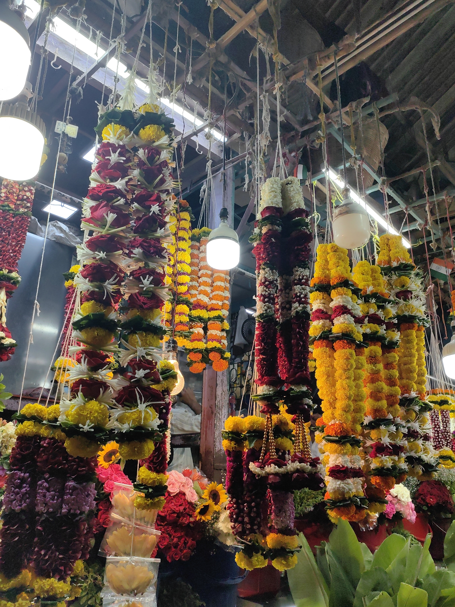 This image depicts a vibrant flower stall, likely in a market in Mumbai, India, given the common practice of selling flower garlands for religious and decorative purposes. The stall is densely packed with various floral arrangements, primarily garlands, hanging from an overhead structure. Dominating the foreground are several long, vertical garlands composed of marigolds in shades of yellow and orange, interspersed with red roses and white flowers. Some garlands also feature gold beads. To the left, darker, richer colored garlands, possibly made of deep red or purple flowers, are visible. The lighting suggests an indoor or covered market setting, with artificial lights illuminating the scene. The background is cluttered, hinting at other stalls or market structures, and a person can be partially seen in the background, suggesting the stall is actively operating. The overall impression is one of abundance and tradition, characteristic of Indian flower markets.