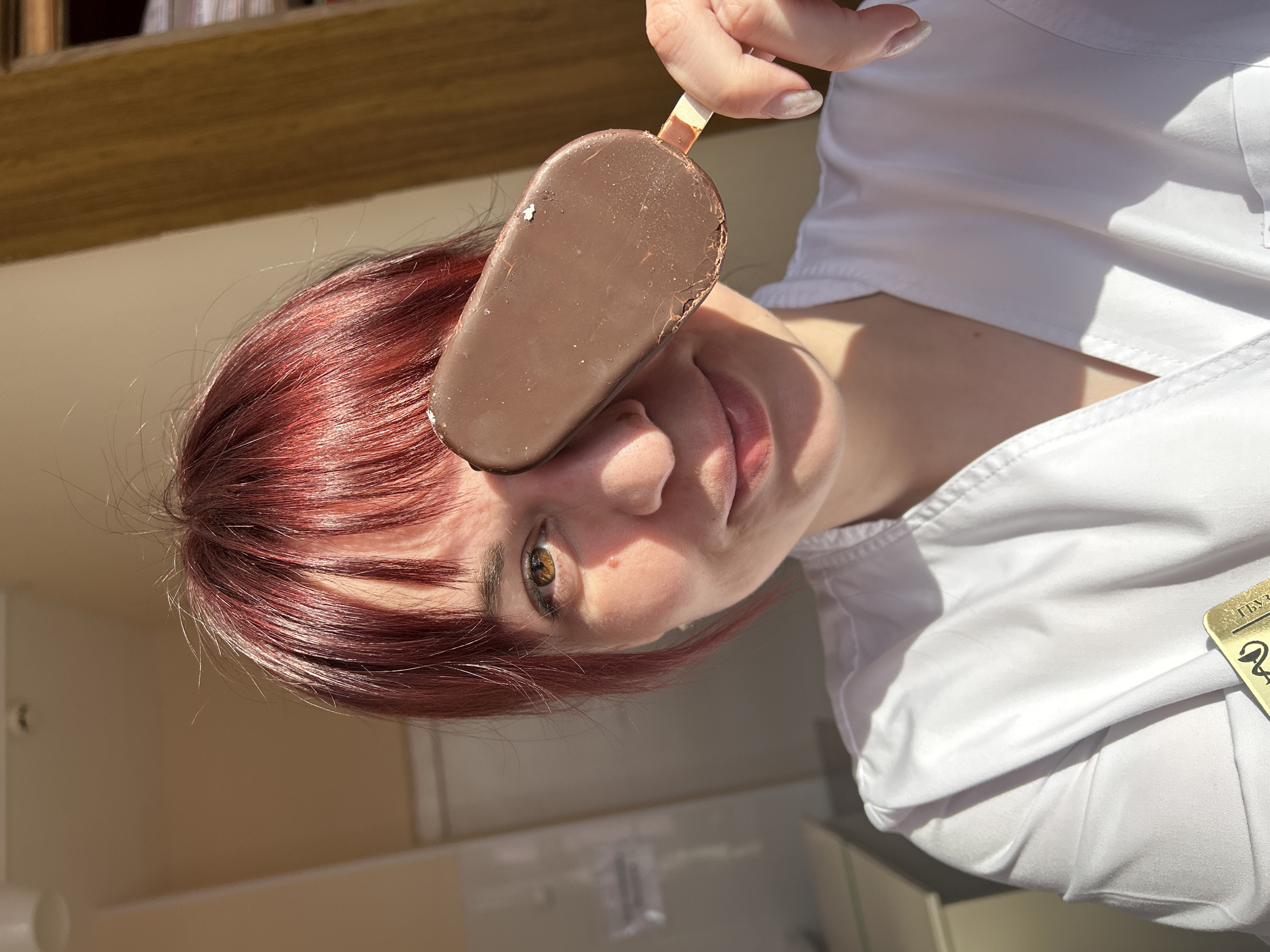 A person with red hair is holding a chocolate-covered ice cream bar in front of their face, covering one eye. They are wearing a white shirt. The background is blurry, but there appear to be shelves or a wooden structure behind them. The location, Фоки, Russia, is not discernible from the image.