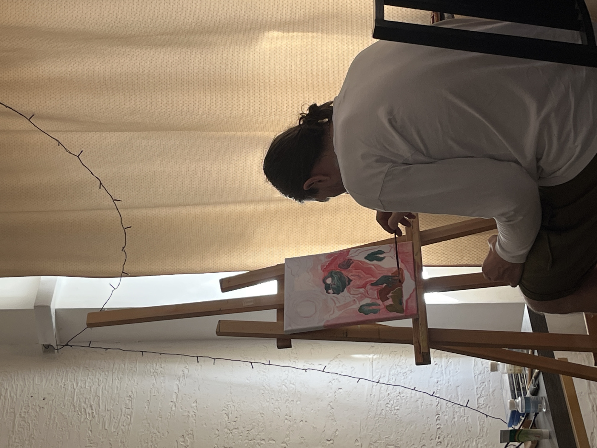 A person with long dark hair in a bun, wearing a white long-sleeved shirt and olive shorts, is standing in front of an easel and painting a canvas. The painting on the canvas is abstract, featuring shades of pink, white, and green. The easel is wooden and positioned on a textured white wall with a string of lights running along it. Above the easel, there's a beige-colored curtain. Near the easel, there are painting supplies. The location is not identifiable as Krasnoyarsk, Russia, based on the image alone.
