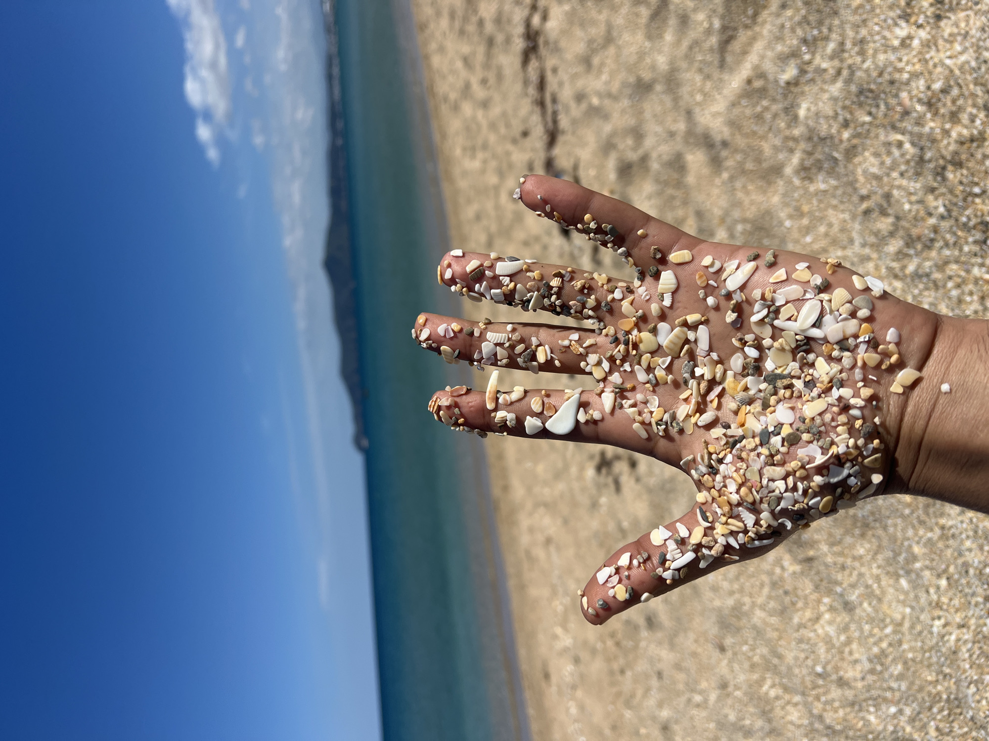 A hand is covered in small shells and pebbles on a beach in M'Diq, Morocco. The sky and sea are visible in the background. The background on the right side of the hand shows the beach extending away from the frame.