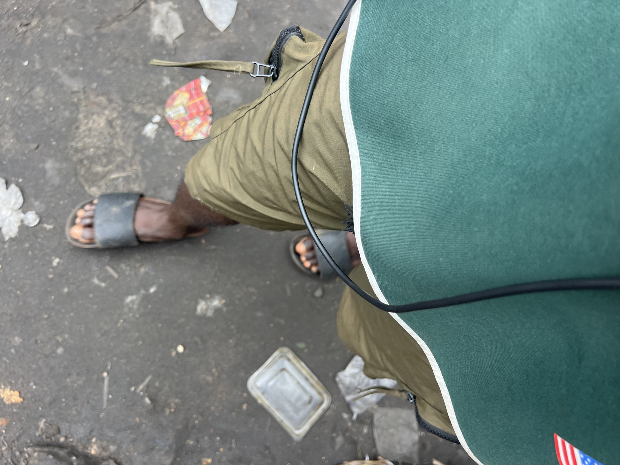 An overhead shot captures the lower body of a person walking on a dark, possibly wet or muddy, paved surface in Nkpor, Nigeria. The individual wears an olive green pair of shorts or trousers, and a dark green top with a white trim along the side is partially visible on the right. A black cable runs diagonally across the green top and over the olive-colored clothing. A black strap with a silver metallic buckle is seen near the left hip, likely part of a bag or accessory.
The person is wearing black slide sandals on both feet, revealing dark skin. The ground is scattered with various pieces of litter, including crumpled white plastic, a red and yellow wrapper that is difficult to decipher, and a clear, rectangular plastic food container with a lid. Other small, indistinguishable debris is also visible on the grimy surface. In the lower right corner, a small portion of what appears to be an American flag patch, with red and white stripes and blue with white stars, is visible on the person's clothing or an attached item. The overall scene suggests an outdoor, urban environment.