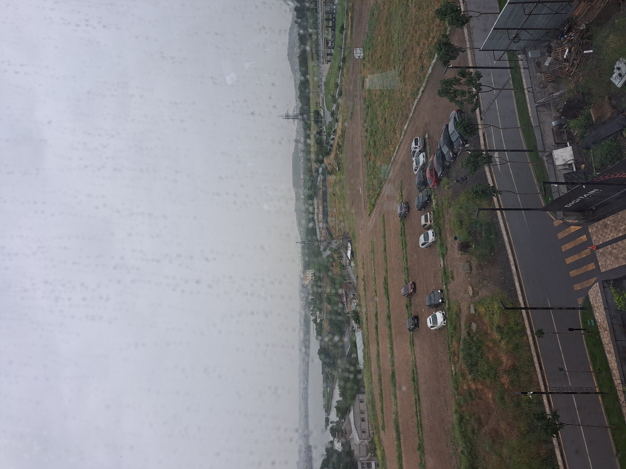 The image shows an aerial view of a landscape taken through a window covered with raindrops. On the left side, there is a body of water, possibly a river or a lake, alongside buildings and vegetation. Adjacent to this area is a long stretch of land with cars parked along the edge of a road, with lamp posts lining the roadside. There appears to be some sort of building development with construction materials visible on the right side of the image. The overall view is of an urban-meets-natural environment on a rainy day, suggesting the outskirts or developing area of Yerevan, Armenia.
