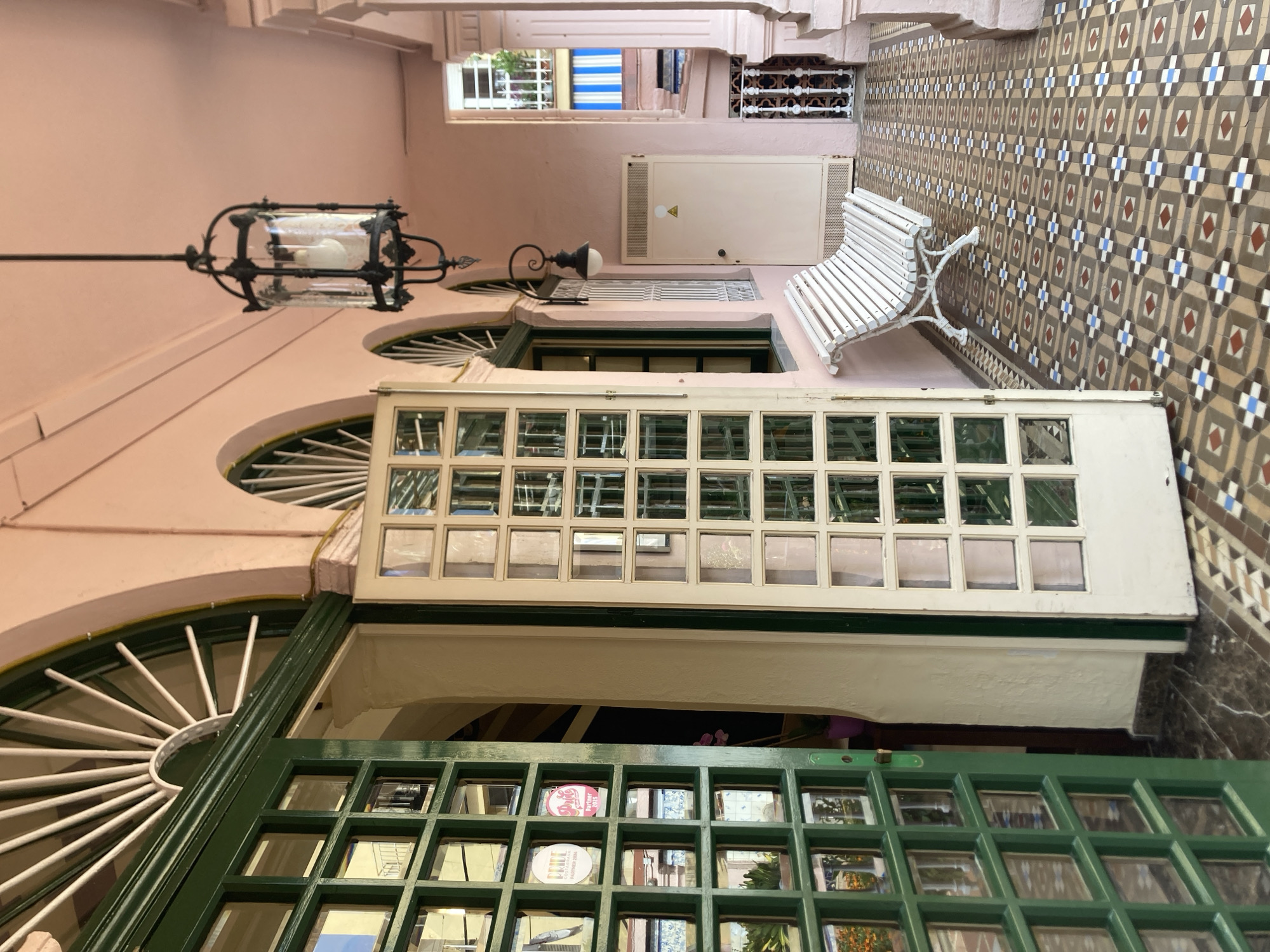 The image depicts a covered walkway or veranda outside a building with a pink facade. The walkway is paved with colorful, patterned tiles. To the left, a tall, dark green double door with many glass panes is partially open, revealing a glimpse of an interior with more glass-paned doors and a hint of plants. To the right, a white slatted bench sits against the pink wall. Above the bench, a vintage-style black lantern hangs from the ceiling. Further down the walkway, a cream-colored utility box with a triangular symbol is visible. In the background, an open-air structure with balconies and blue and white striped awnings is visible. The architecture suggests a Mediterranean or colonial style. The scene appears to be located in Lloret de Mar, Spain, an area known for its vibrant coastal architecture and historic buildings.