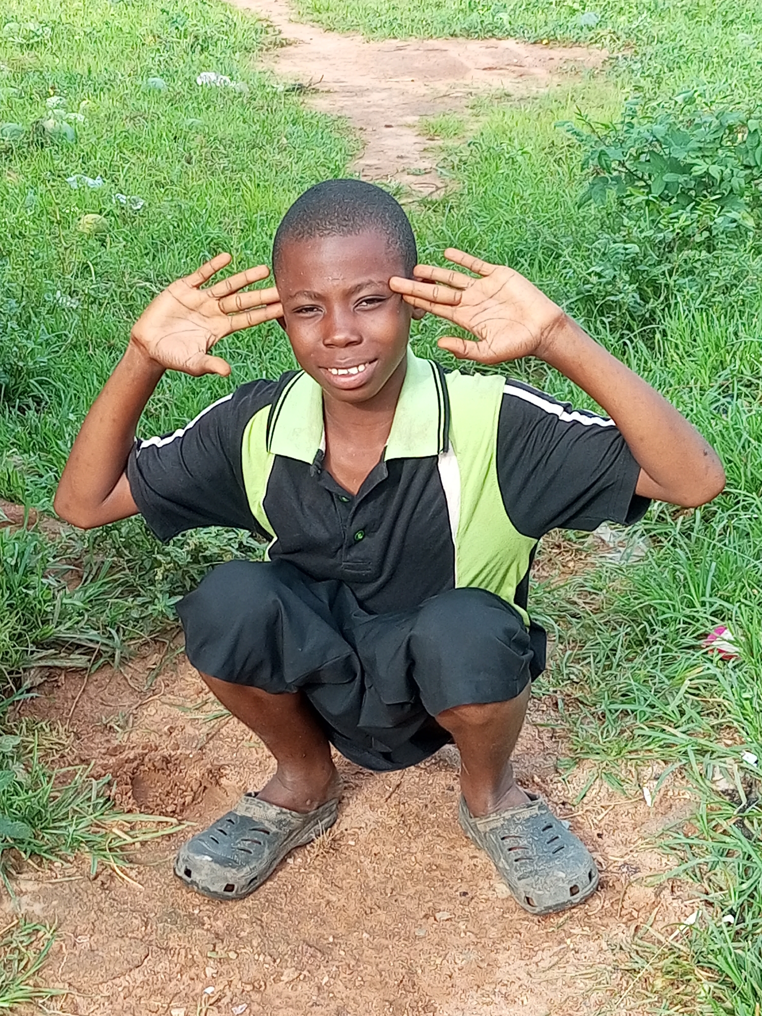 A young boy crouches in dirt with grass surrounding him and a path behind him. He wears a black t-shirt with green and white accents on the collar and sleeves, black shorts, and dirty grey clogs. He is smiling and has his hands raised to the sides of his head, palms facing forward. The scene appears to be outdoors, in a rural or natural setting, possibly in Tempure, Nigeria, given the context. The lighting suggests it is daytime, and the greenery indicates favorable weather for outdoor activity. The boy's expression and pose convey a playful and cheerful emotion. There is no visible text in the image.
