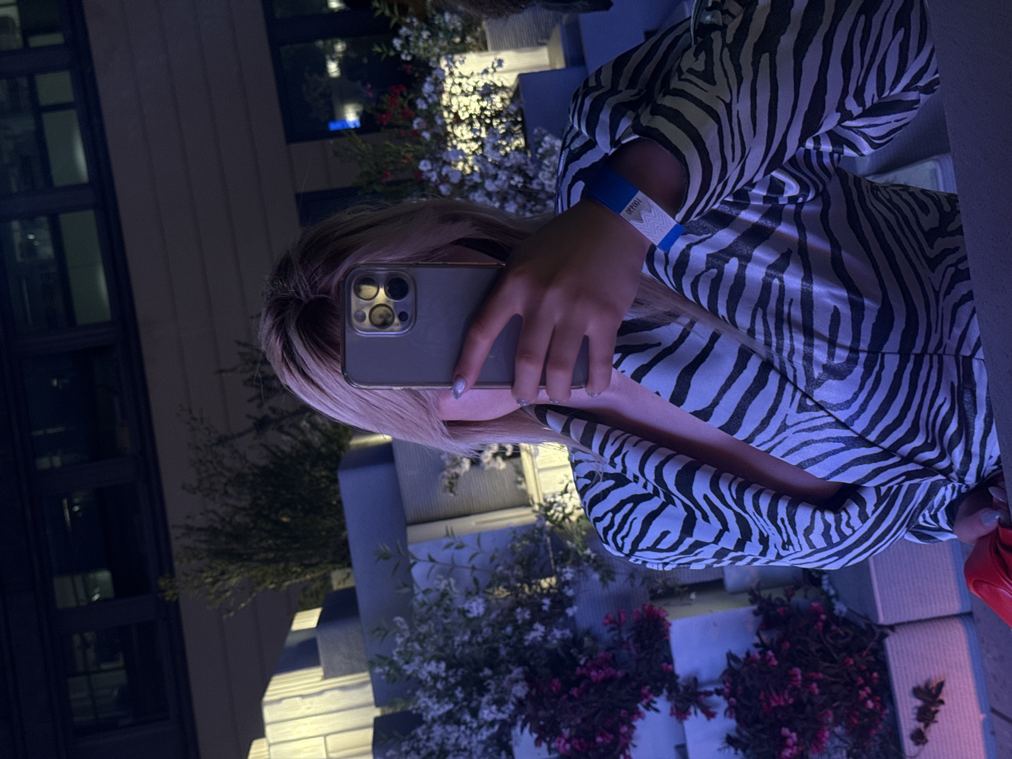 A person with blonde hair is taking a picture of themself with a smartphone. The person is wearing a zebra-striped top and a blue wristband. The location appears to be outdoors, possibly in a courtyard in Milan, Italy, considering the architectural style and the presence of flowering plants.