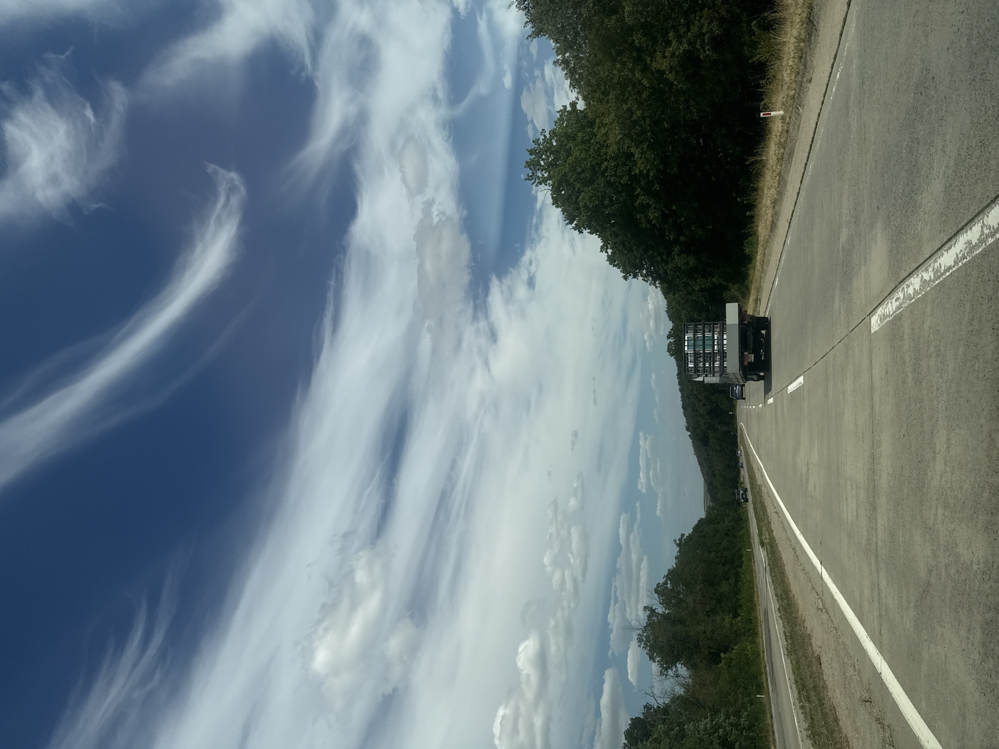 The image shows a view from a vehicle traveling on a highway. The road is made of concrete and has white lane markings. In the foreground, the road surface is visible. In the middle ground, a truck carrying what appears to be stacked crates is traveling in the same direction. Several other vehicles are visible further down the road, including cars. The sides of the highway are lined with trees and vegetation, with rolling hills in the background. The sky is a deep blue with wispy white clouds. The image appears to be taken during the daytime, with ample natural light. The location is the highway near the city of Răzeni, Moldova.