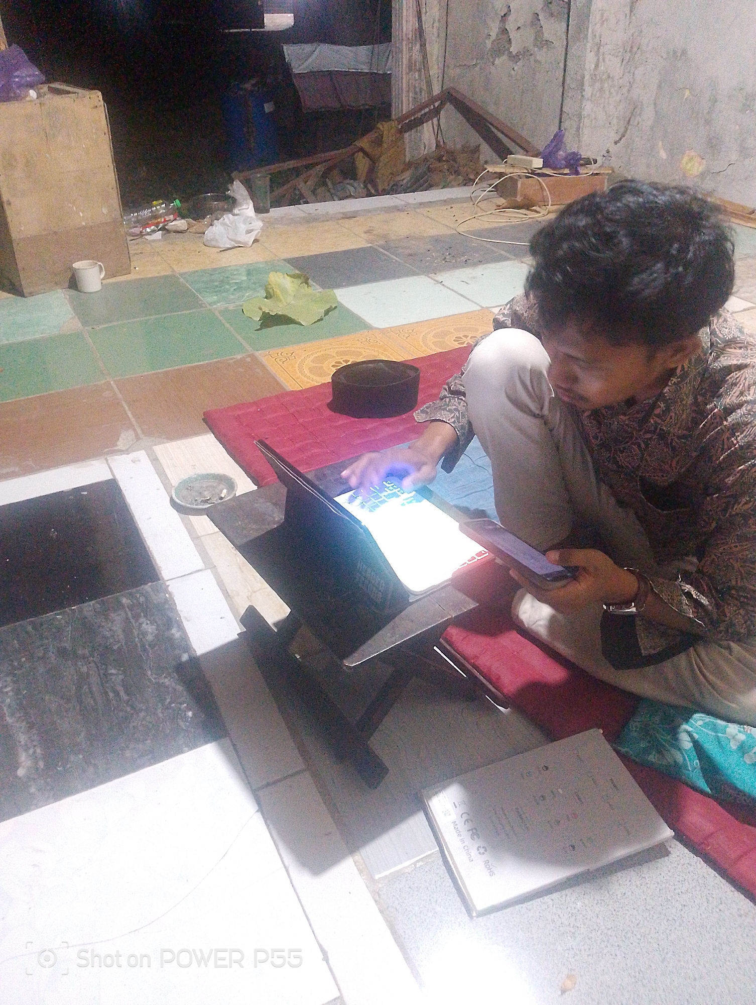 A person is sitting on the floor in what appears to be an outdoor or semi-outdoor setting, possibly a patio or courtyard. The ground is tiled with a pattern of squares in varying colors including white, black, green, and brown. A large, light brown wooden box is positioned to the left of the frame. In front of the person, a laptop is open and placed on a dark stand. The person is looking down at the laptop and holding a smartphone in their right hand. The person is wearing a patterned long-sleeved shirt and light-colored pants. A dark, rounded cap is visible to the right of the laptop. There are various items scattered around, including crumpled white plastic, a plastic bottle with a green cap, and a small ashtray with cigarette butts. In the background, there is debris, some wooden structures, and a concrete wall with peeling paint. The lighting suggests it is nighttime or dusk. A watermark at the bottom left of the image reads "Shot on POWER P55".