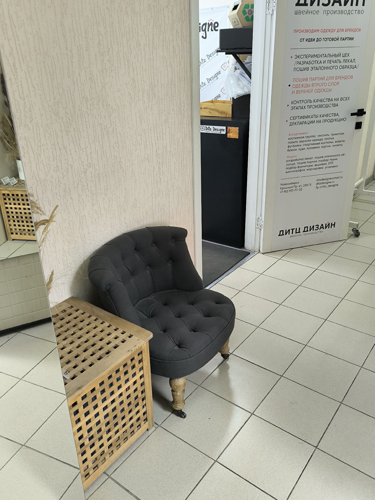 The image shows a retail or workshop space with a focus on a sign for "ДИТЦ ДИЗАЙН" (DITZ DESIGN), a sewing production company. The sign lists services such as garment production for brands, pattern making, and quality control. Contact information for Novosibirsk, Russia is provided, including a phone number and email address.
In the foreground, a dark upholstered armchair with tufted detailing is positioned next to a light-colored wooden storage unit with a lattice design. The floor is tiled with a grid pattern. To the left, a portion of a large mirror reflects the environment, including dried grasses in a vase and parts of the wooden storage unit. The wall behind the armchair and storage unit has a textured, light-colored finish. To the right of the armchair, the doorway reveals more of the interior space, including a black cabinet and part of a black mat on the floor. The overall scene suggests a business establishment with a waiting area or display space.