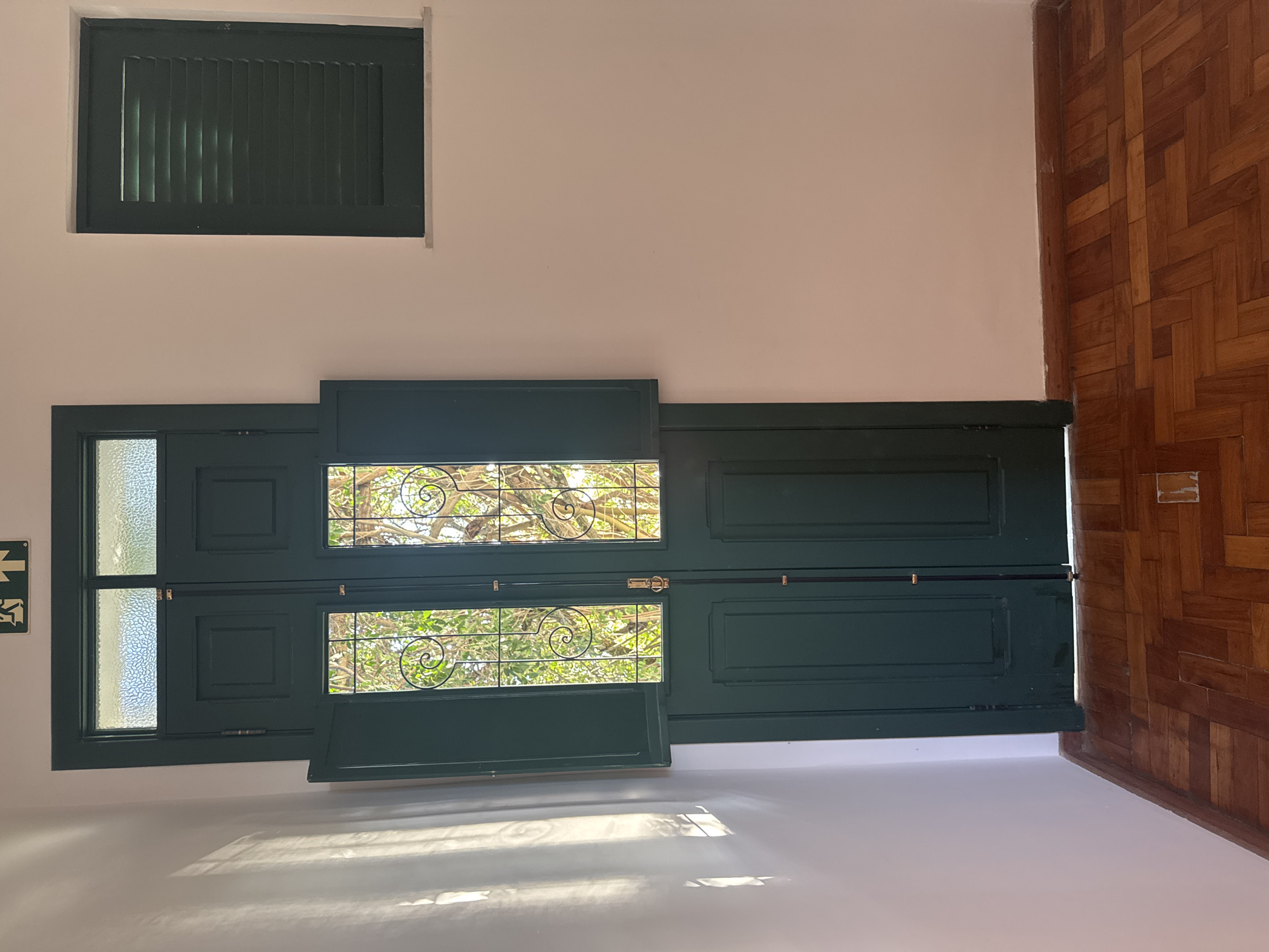 The image captures an interior scene featuring a dark green double door with open shutters, revealing decorative metalwork and foliage. Above the door, there are two frosted glass panels and an illuminated green exit sign. To the right of the door, a dark green louvered window is visible on a white wall. The floor is made of polished wooden parquet. The lighting suggests daytime, with sunlight casting diagonal shadows on the left wall. No people or animals are present in the scene. The location is identified as a residential building in Niterói, Brazil.