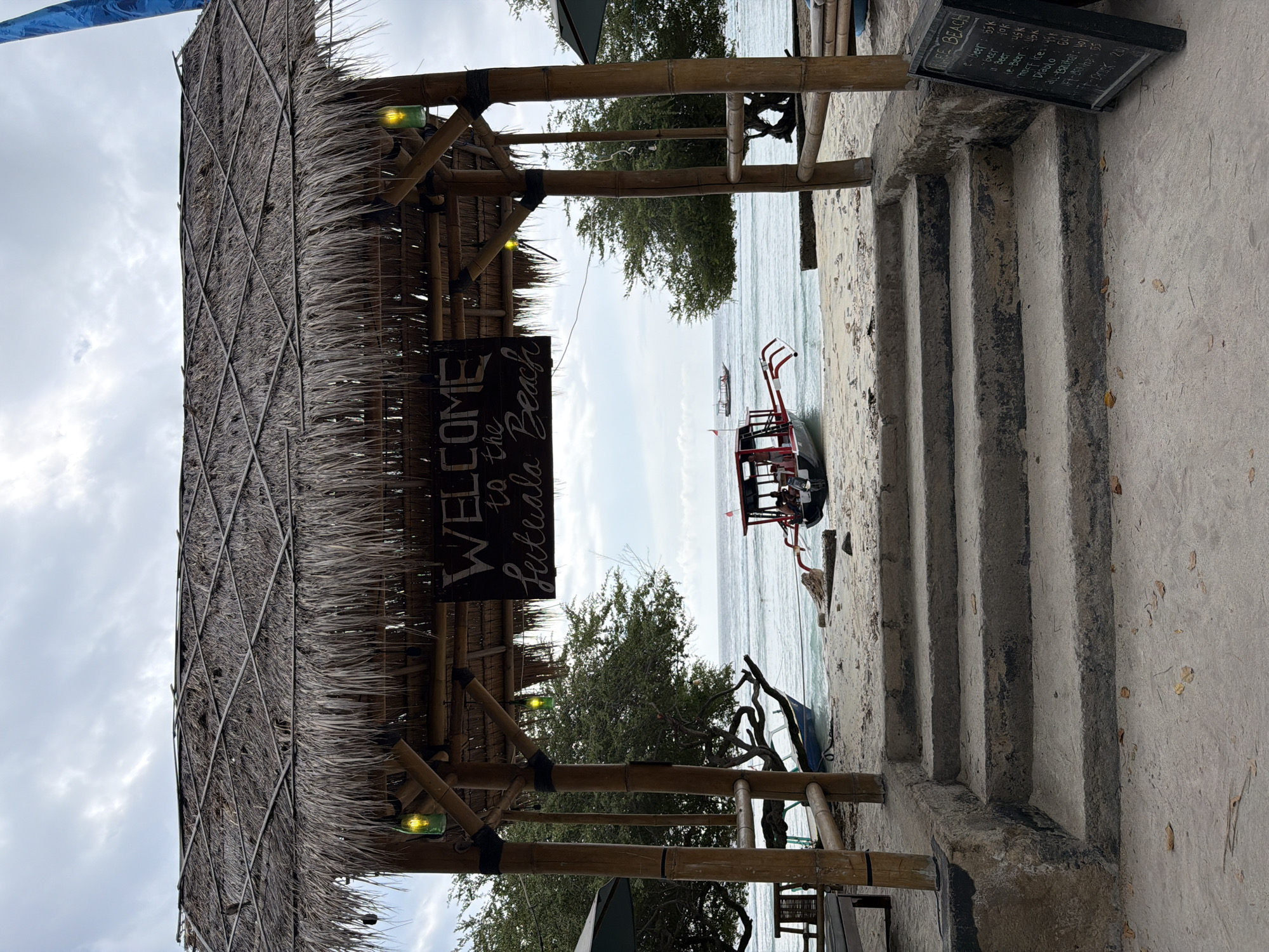 The image depicts a view of a beach in Unknown, Indonesia, framed by a thatched-roof structure. The structure has bamboo supports and is adorned with two green lights. A sign hanging from the structure reads "WELCOME to the Lutuala Beach" in white lettering. In front of the structure are concrete stairs leading down to a sandy beach. The beach has several boats, including a traditional outrigger boat with a red and white hull. In the background, the ocean stretches to the horizon under a cloudy sky. A chalkboard menu is visible to the right of the stairs. The scene appears peaceful and inviting, typical of a tropical beach destination.