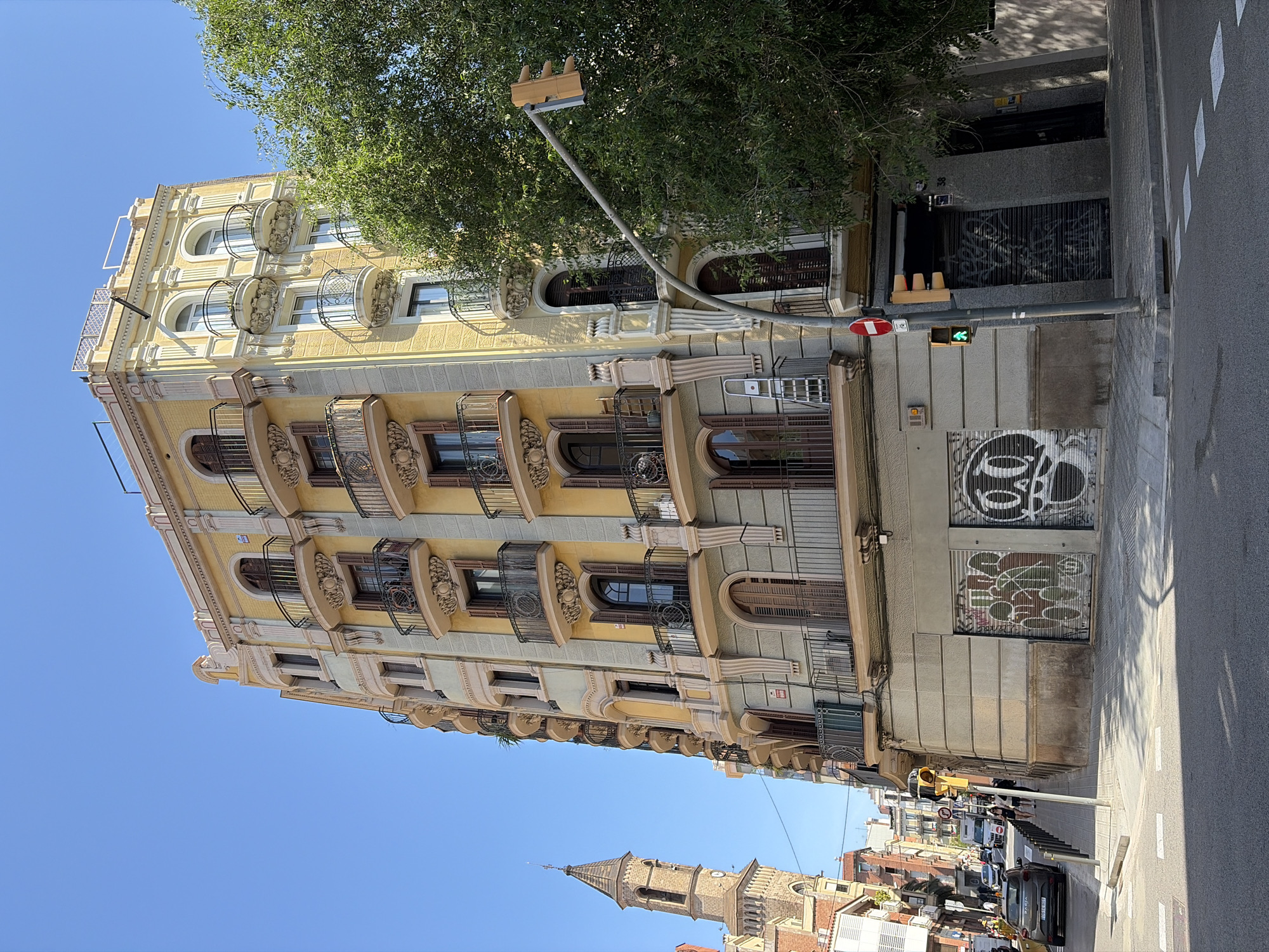 The image shows a street scene in Barcelona, Spain. A tall, ornate building with yellow and beige facades dominates the left side of the frame. The building features decorative balconies and arched windows. A street lamp extends into the frame, partially obscured by the leaves of a tree. Graffiti is visible on the ground floor of the building.
In the background, a church tower rises above the surrounding buildings. Cars and other buildings can be seen further down the street. A pedestrian is visible on the sidewalk. The sky is clear and blue.