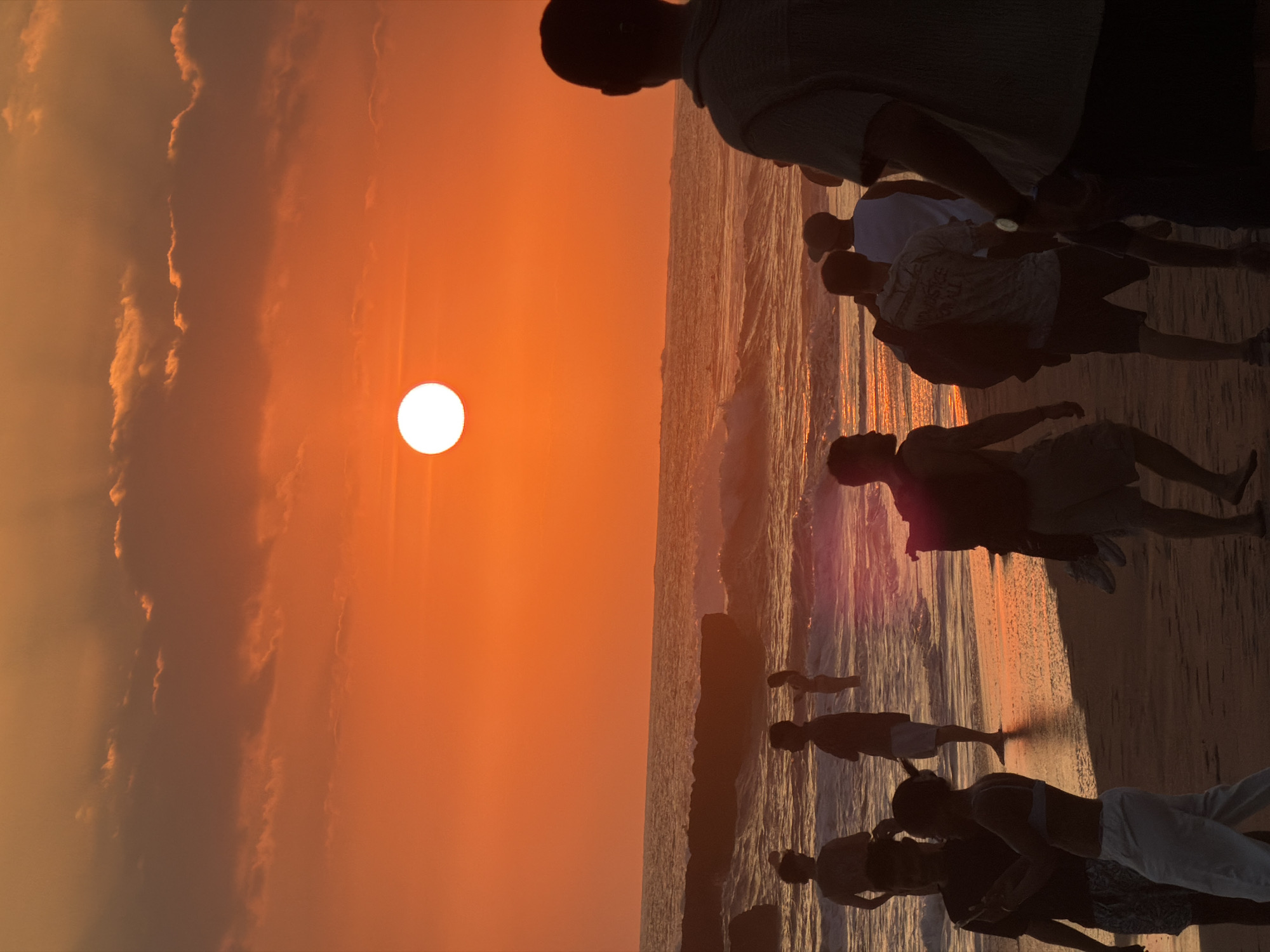 The image depicts a beach scene at sunset in North Kuta, Indonesia. The sky is a vibrant orange with diffused sunlight, and the sun itself is a bright, white orb visible above the horizon. The ocean has moderate waves, and the water reflects the warm hues of the sky. Several people are silhouetted on the sandy beach and in the shallow water. On the beach, individuals are standing and walking, some facing the ocean and others interacting with each other. In the water, a few figures are visible wading. The overall atmosphere is one of leisure and natural beauty during twilight.