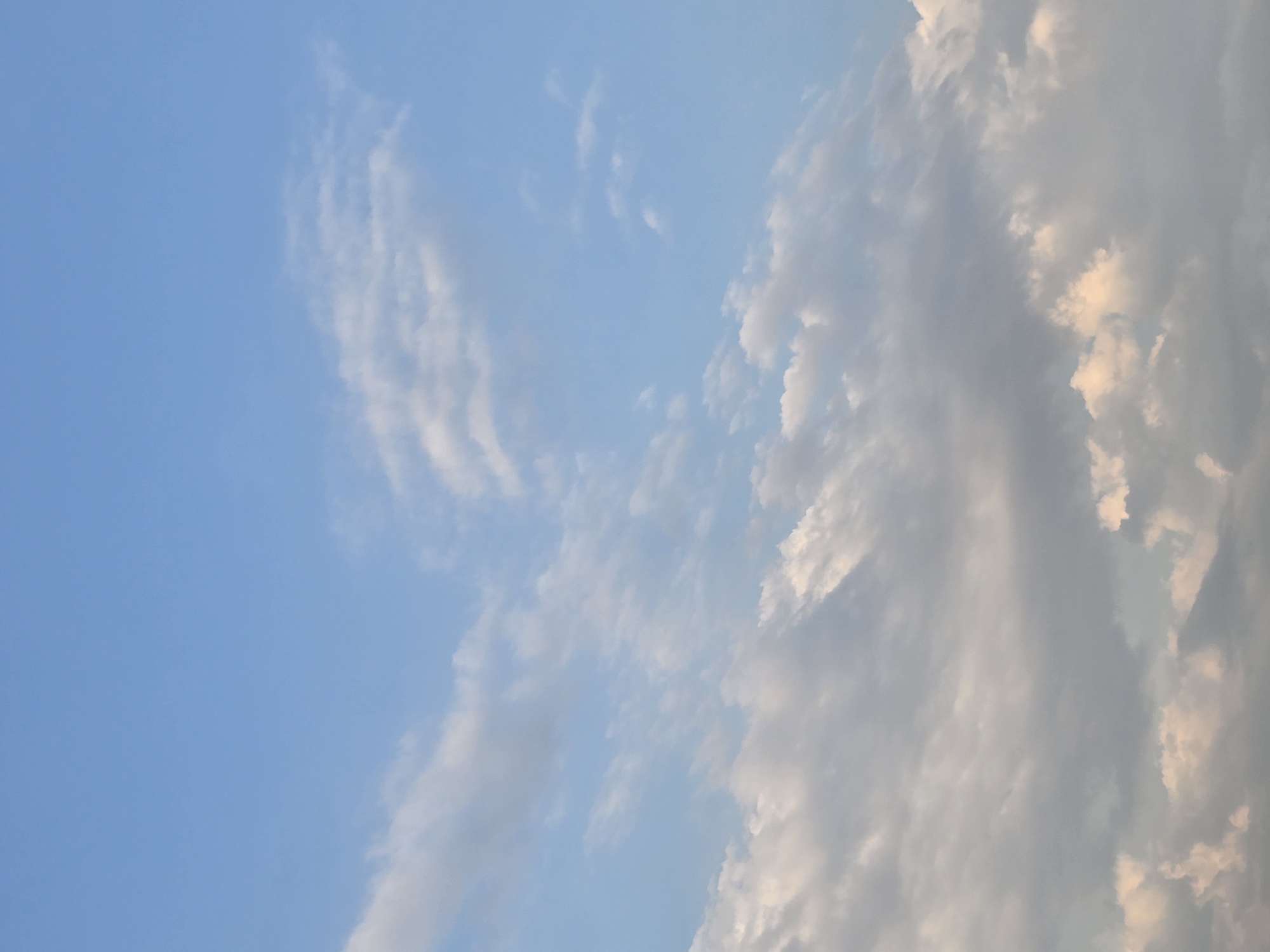 The image shows a clear blue sky with scattered clouds. The clouds are white and fluffy in some areas, while others are darker and more diffused, suggesting varying altitudes and densities. The lighting indicates it is daytime, likely late afternoon or early morning due to the soft glow on some of the clouds. The scene is devoid of any man-made structures or individuals, focusing solely on the natural atmospheric phenomenon. The setting is likely an outdoor environment with an unobstructed view of the sky. There is no information about the location in Medun, Montenegro, within the image itself.
