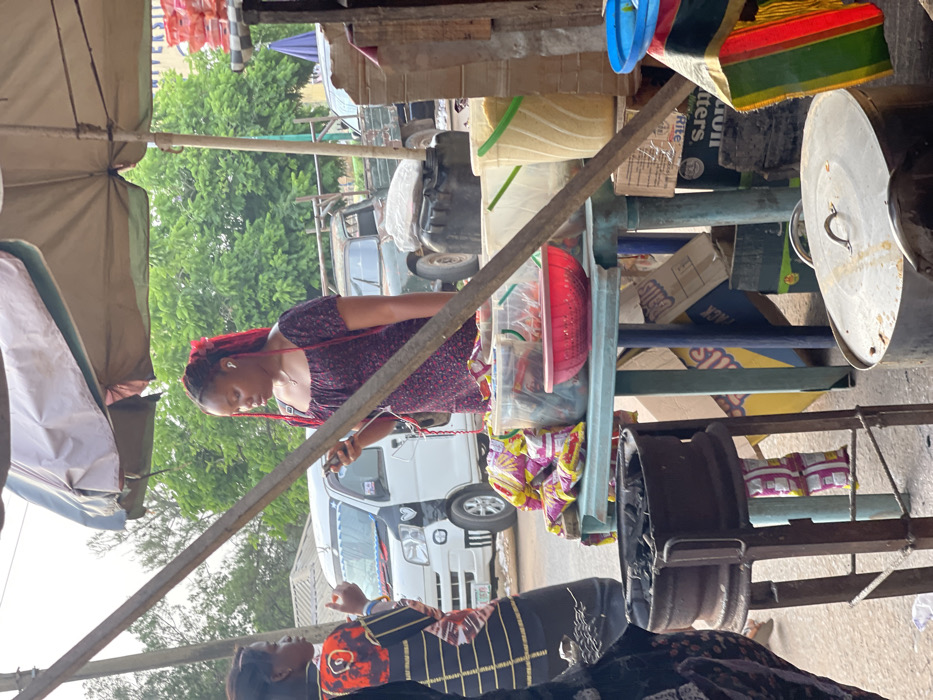 The image captures an outdoor market scene, likely during the daytime, with several people and various goods visible under what appears to be a slightly overcast sky.
In the foreground, a woman with reddish-brown braided hair, possibly dreadlocks or twists, is standing to the left of a makeshift stall. She is wearing a dark, short-sleeved top with a subtle pattern and has white earbuds in her ears. Her gaze is directed downwards, suggesting she might be looking at a phone or an item in her hands.
The stall itself is constructed from metal framing and shelves, laden with various items. On the upper shelves, clear plastic containers hold goods; one contains a red bowl or lid, and another larger container appears to hold light-colored grains or produce. Below these, there are several plastic bags of snacks, identifiable by their pink and white packaging. Cardboard boxes are also stacked on and around the stall, with some partially visible text, including "BUTTERS," "FRUIT," "Dolls" (or "Balls"), and "PACK." A large metal pot with a lid rests on the right side of the stall, and a colorful striped bag (red, yellow, green) hangs near it. A sturdy diagonal beam, possibly part of the stall's support or another nearby structure, crosses the mid-ground.
To the far left, a light-colored fabric canopy or umbrella structure provides shade, supported by visible poles. In the background, several vehicles are parked on what appears to be a road or open area. A white mini-van or small bus with dark decorative accents (possibly heart shapes) on its front is clearly visible behind the woman with red hair. Further back, an older, possibly darker-colored truck or van with a spare tire mounted on the rear is also present. Green foliage from trees is visible above and between the vehicles.
In the bottom right corner, another woman is partially visible, wearing a dark top with orange and black patterns. She appears to be looking upwards or towards the left, possibly interacting with someone or observing the scene.
The overall atmosphere suggests a busy, utilitarian market environment.