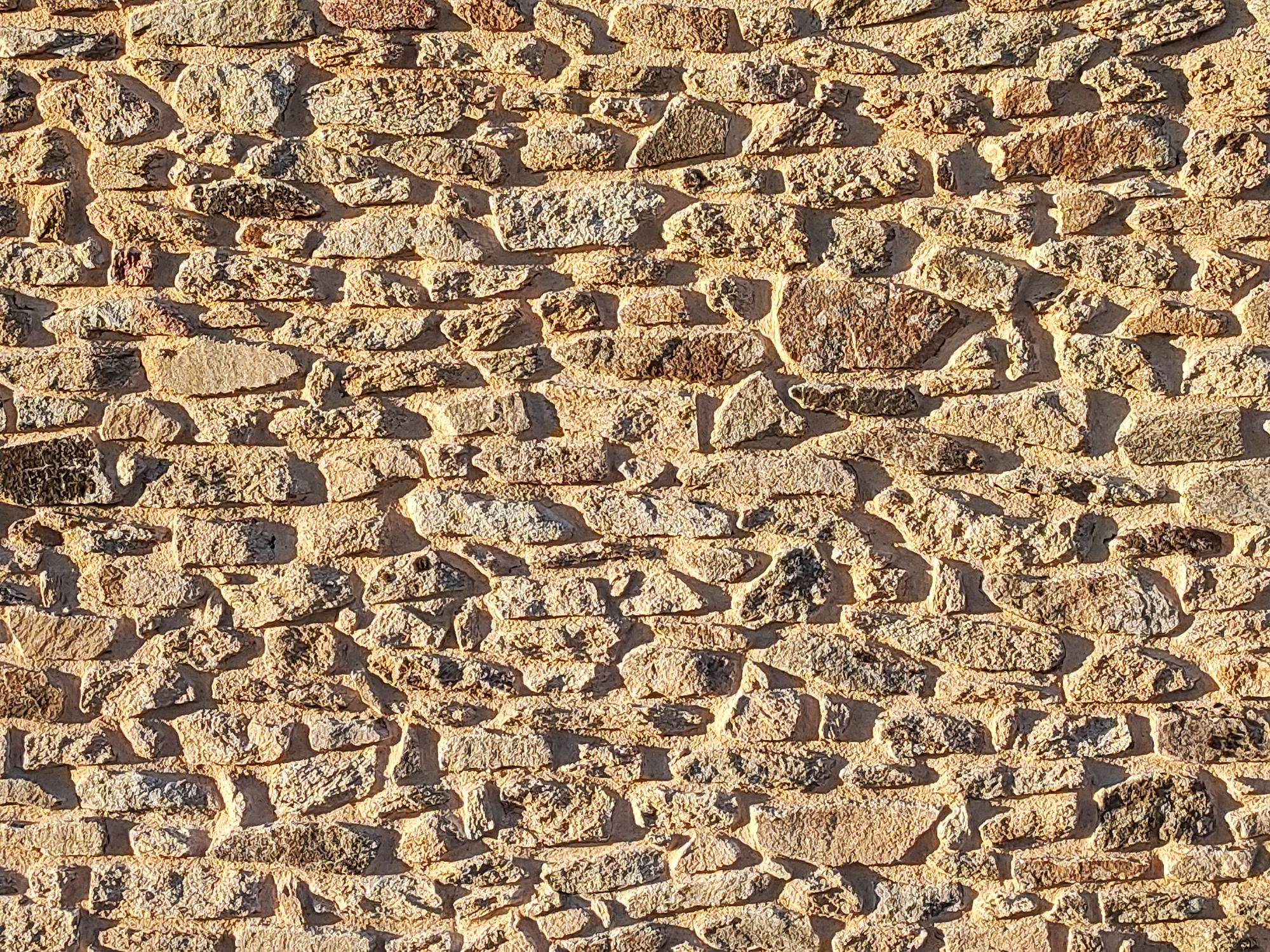 The image is a close-up, full-frame shot of a rustic stone wall. The stones are irregularly shaped and vary in size and color, ranging from light tan and cream to darker browns and grays. They appear to be rough-hewn and naturally shaped, fitted together with mortar. The texture of the stones is uneven, with many showing rough surfaces and chipped edges. The mortar is a lighter, sandy color, and it fills the gaps between the stones, creating a textured, somewhat uneven surface. Sunlight casts strong shadows across the wall, highlighting the three-dimensional texture of the stones and creating a strong contrast between the lit and shadowed areas. The lighting suggests it is daytime, likely with the sun at an angle to the wall. The image captures the detail and texture of the stonework without any other contextual elements. It's a detailed view of a building material, showcasing its natural irregularities and the effect of light and shadow on its surface.