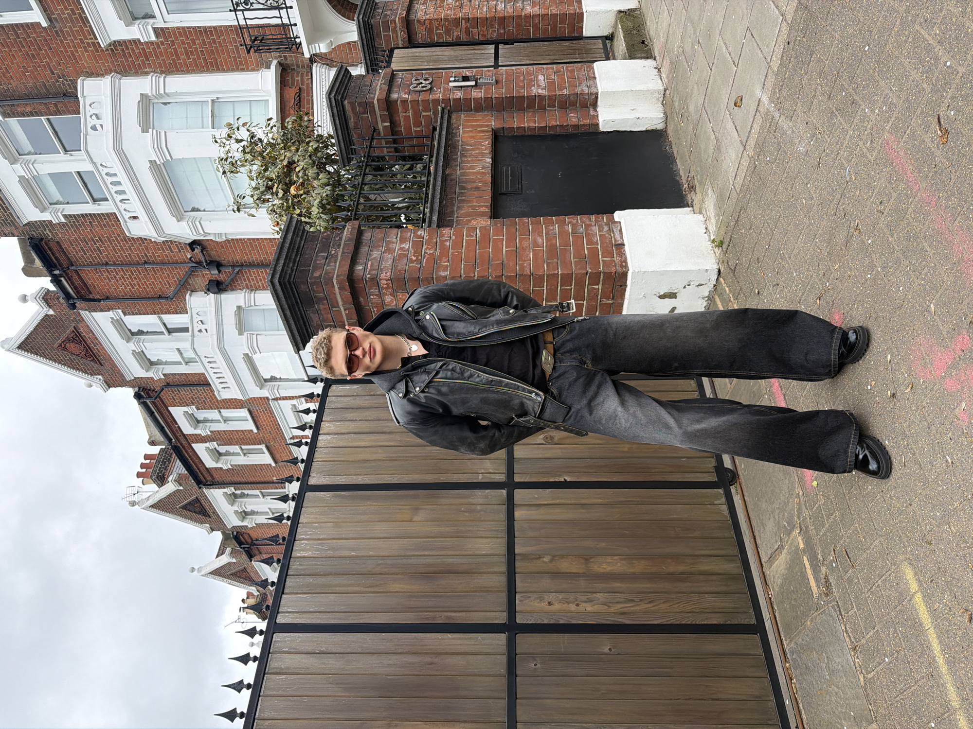 The image depicts a fair-skinned male with short, blond hair and wearing tinted sunglasses. He is dressed in a black leather jacket over a black top, dark gray jeans, and black shoes. He is posed outside on a pavement. Behind him is a low wooden fence with metal accents and brick townhouses with ornate white trim. The buildings feature prominent brick facades, multiple windows, and some have black iron railings. The paving stones are gray and there are hints of pink paint markings. The location is likely a residential street in London, United Kingdom.