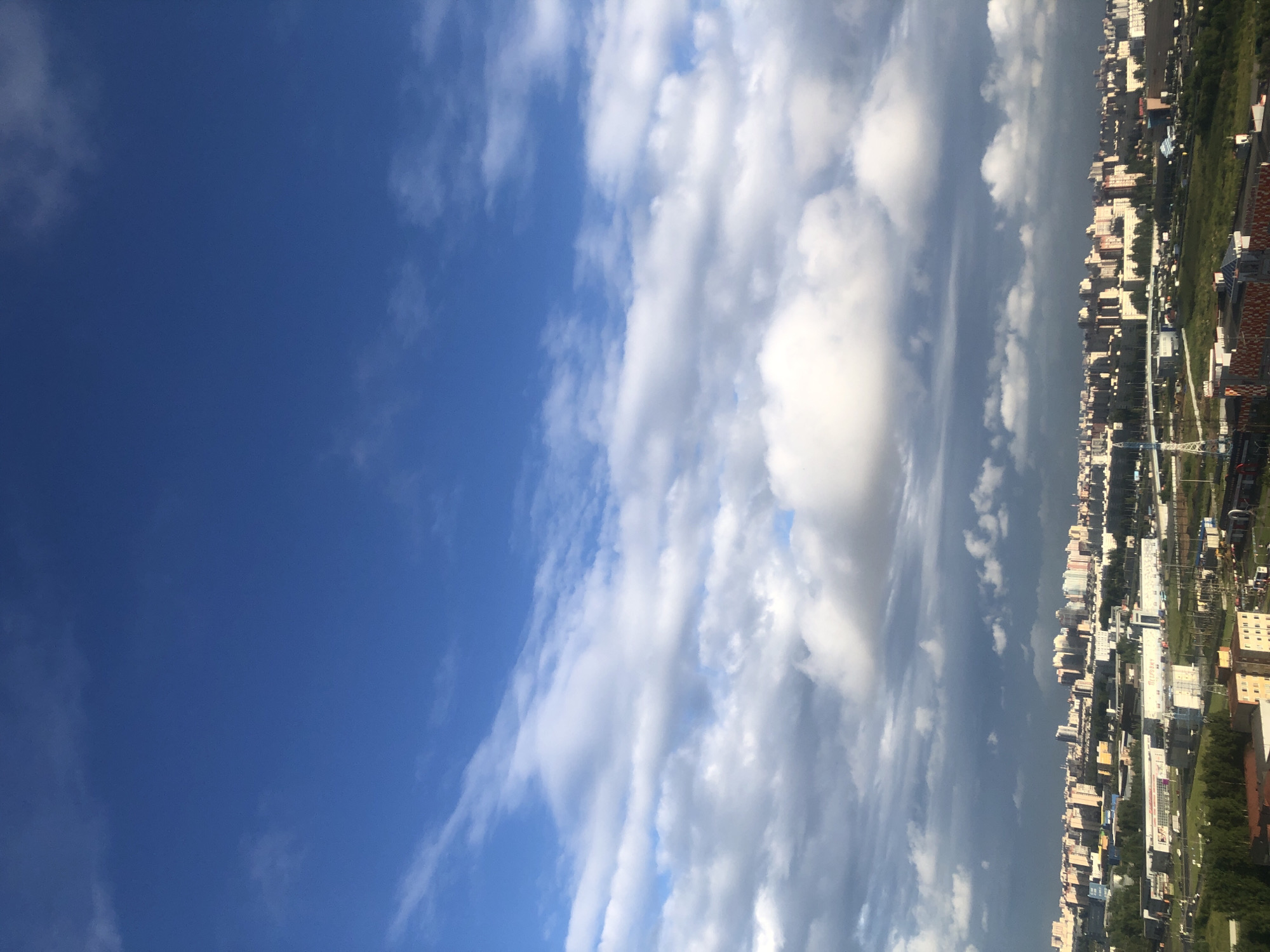 This media file is a photograph taken from a high vantage point, possibly an airplane or a tall building. The image is oriented vertically, with the left side dominated by a clear blue sky with scattered white clouds. As the view moves to the right, a city skyline emerges. The city appears to be densely populated with numerous apartment buildings of varying heights and architectural styles, some with visible balconies and windows. The buildings are a mix of light-colored facades, with some displaying yellow, beige, and white colors.
Interspersed with the buildings are areas of green foliage, suggesting trees and parks. Infrastructure elements like roads, power lines, and what appear to be construction cranes are also visible within the urban landscape. A few signs with text are present on some of the buildings, but they are not clearly legible due to the distance and image resolution.
The weather appears to be fair with a mix of bright sunshine and some cloud cover, casting shadows on parts of the city. The time of day seems to be late morning or early afternoon, judging by the lighting conditions. The overall impression is of a modern, sprawling city under a partly cloudy sky. The image does not contain any people.