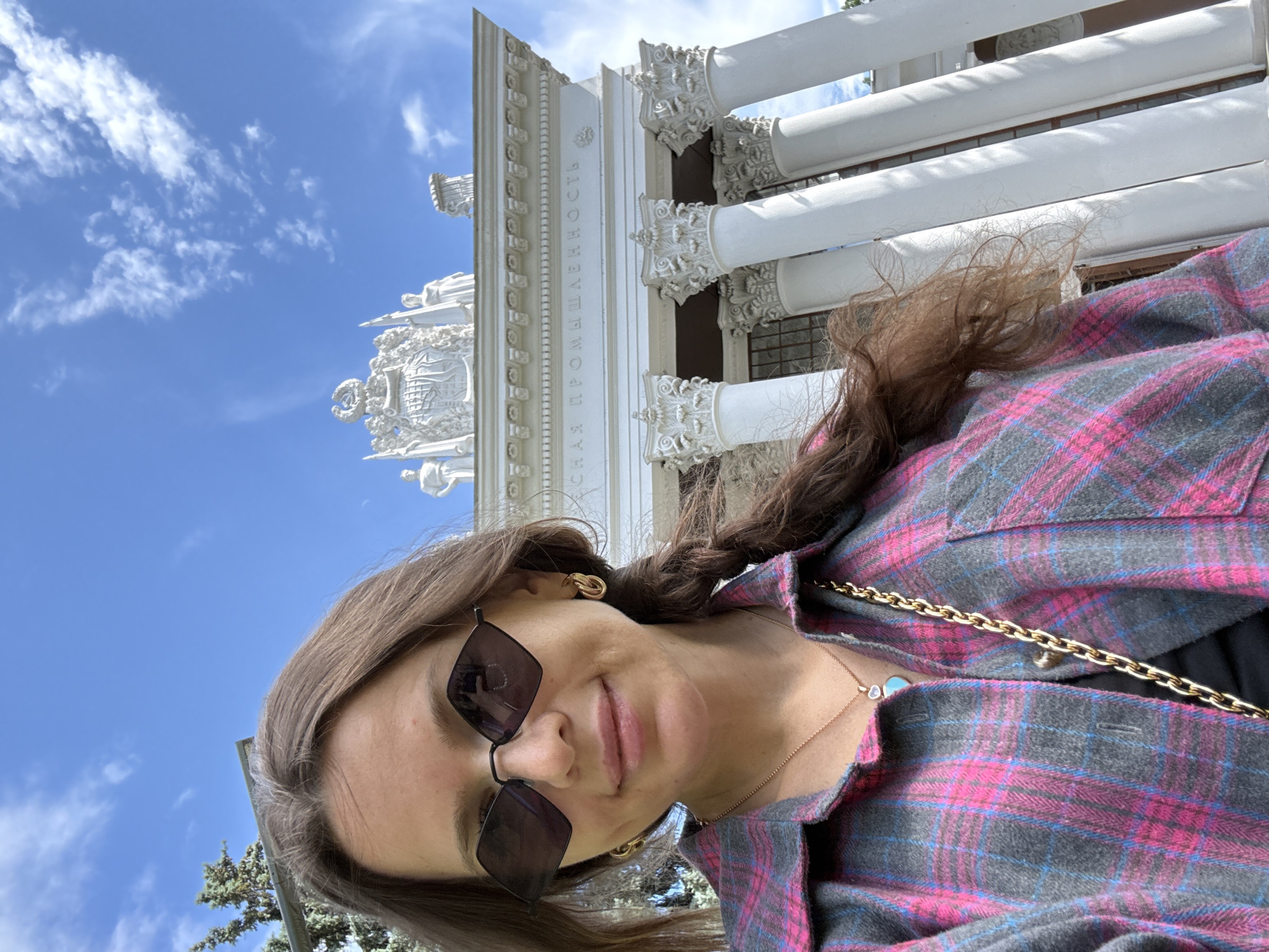 A woman takes a selfie outdoors in front of a white, ornate building. She is wearing a pink and grey plaid shirt, sunglasses, and a gold necklace. The building appears to be historic, with large columns and intricate carvings. The sky is blue with scattered white clouds, suggesting it is daytime and the weather is pleasant. The overall scene conveys a sense of sightseeing or tourism, possibly at an exhibition center or museum given the architectural style. Visible text on the building, in Russian, appears to read "ВСЕРОССИЙСКАЯ ПРОМЫШЛЕННОСТЬ", which translates to "ALL-RUSSIAN INDUSTRY".