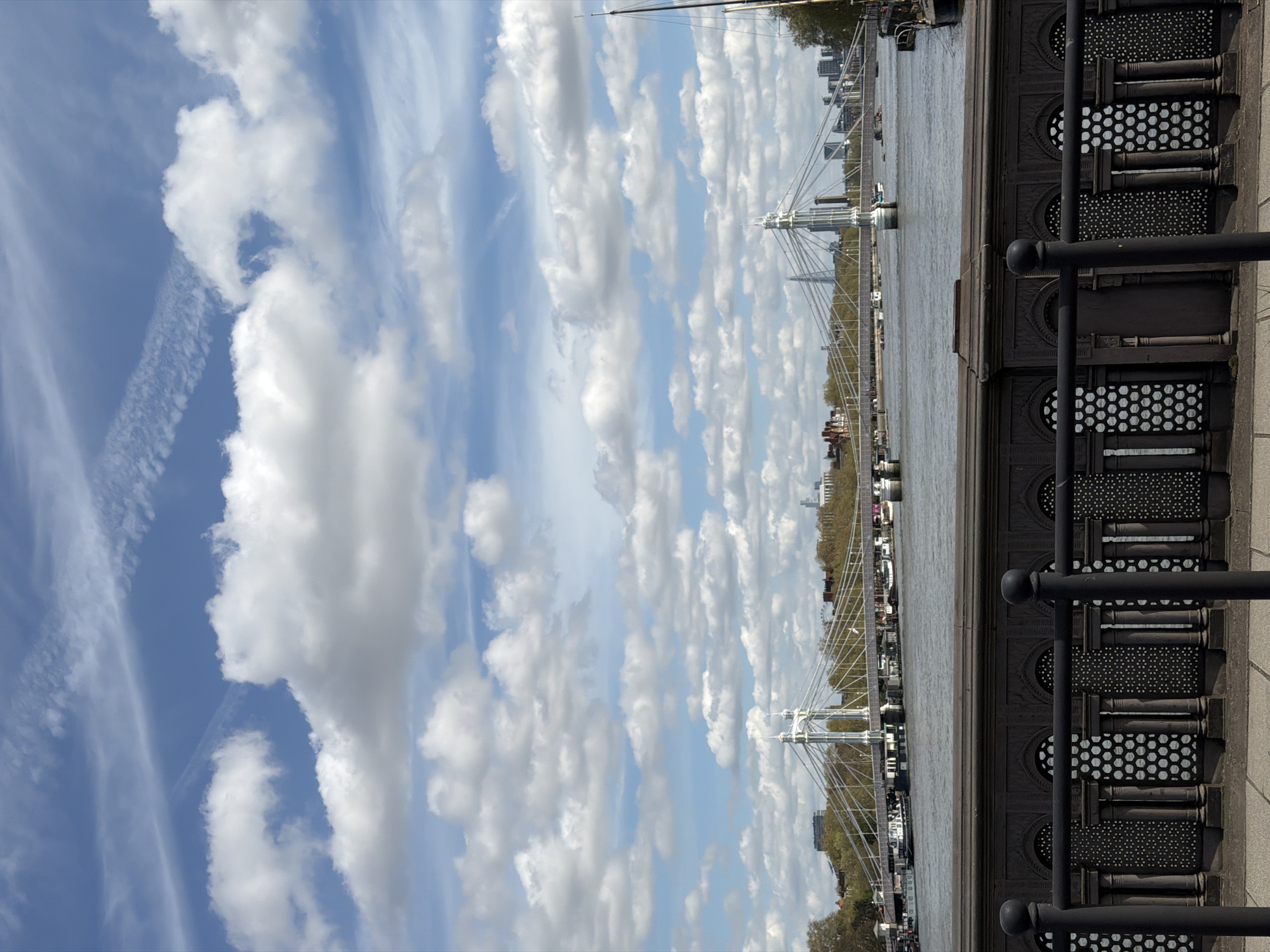 The image shows a view of the Hungerford Bridge and Golden Jubilee Bridges in London, UK. The perspective is from a position on the embankment, with a decorative railing visible in the foreground. The Thames River is in the middle ground, with the bridges stretching across it. The sky is predominantly blue with scattered cumulus clouds. Beyond the bridges, buildings are visible on the opposite bank of the river.