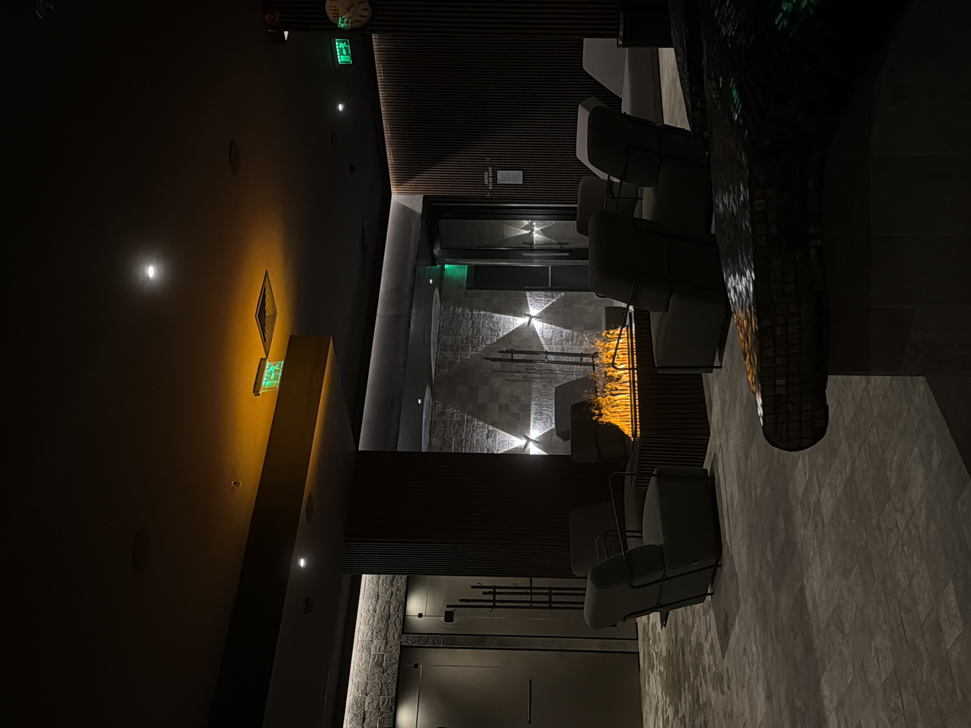 The image shows an indoor seating area with a modern fireplace as the central focus. The room is dimly lit, with warm lighting accentuating the flames and parts of the ceiling. Around the fireplace, several upholstered armchairs are arranged. In the background, there are large, textured walls and doors, suggesting a spa or relaxation facility. There is a clock on the right wall and exit signs visible on the ceiling. The floor is tiled with a geometric pattern. The overall atmosphere is serene and luxurious. The location is identified as a spa within a hotel in Sofia, Bulgaria. No people are visible in the image.