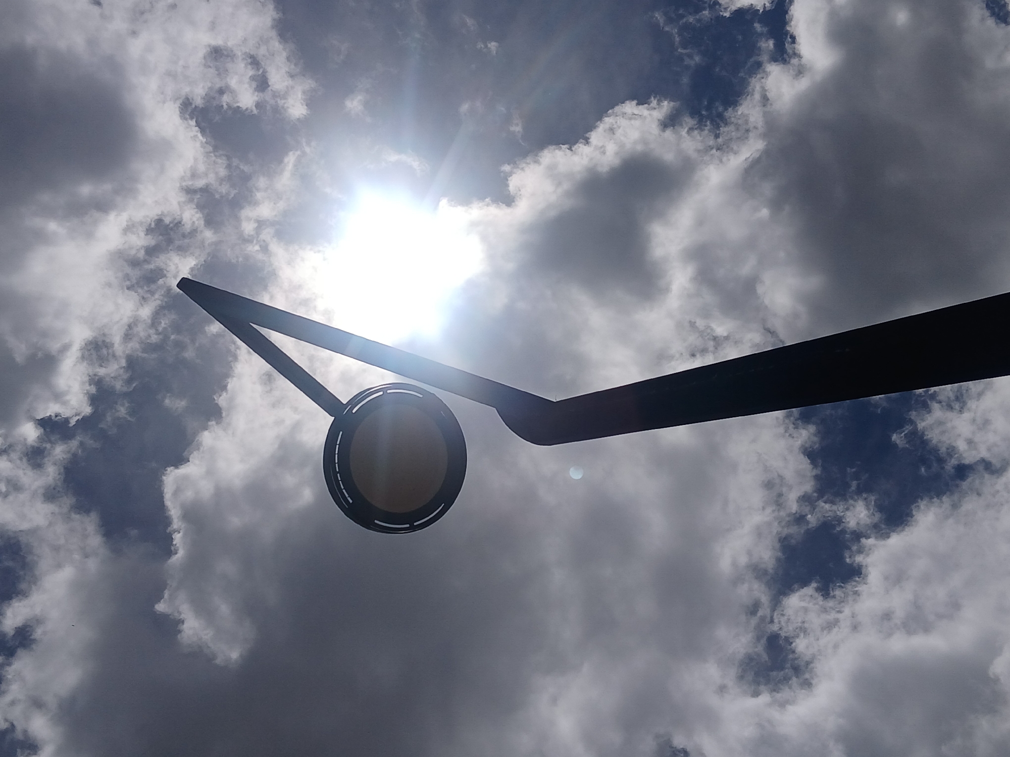 A low-angle shot looking upward shows a round street lamp with a gray-yellow center, set against a cloudy sky with the sun shining brightly through the clouds. The clouds range from white to dark gray, with patches of blue sky visible. The lamp is suspended from a dark, angular support structure. The location cannot be determined as Ciudad Guayana, Venezuela from the content of the image.