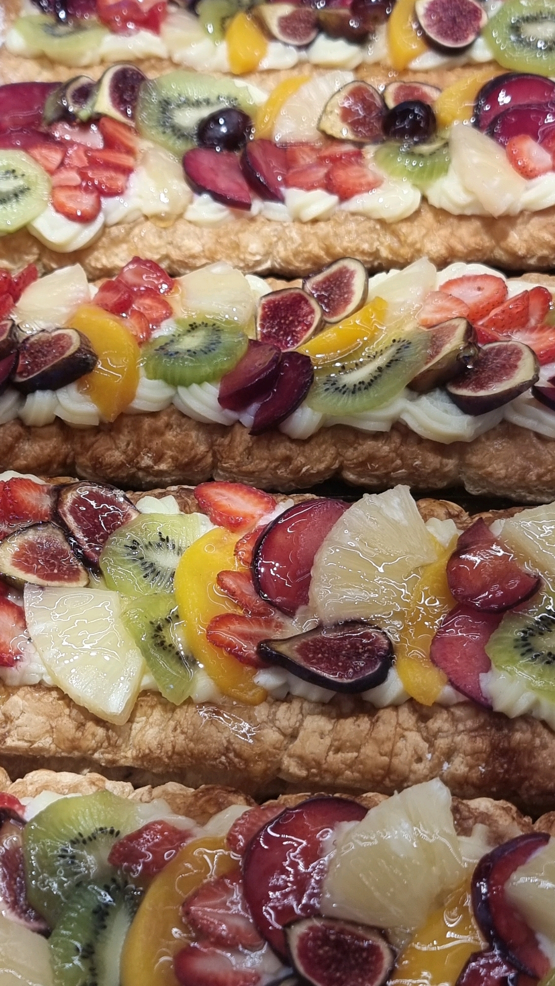 The image displays three rows of fruit tarts, each adorned with an assortment of colorful fruits and a white cream filling. The fruits include sliced strawberries, kiwi, figs, plums, and possibly peaches or apricots, all glazed to a glossy finish. The tarts are layered on a crispy, golden-brown pastry base. The perspective is a close-up, overhead view of the tarts, suggesting they are laid out on a flat surface, possibly in a bakery or display case. There is no discernible environment or urban context within the image itself.