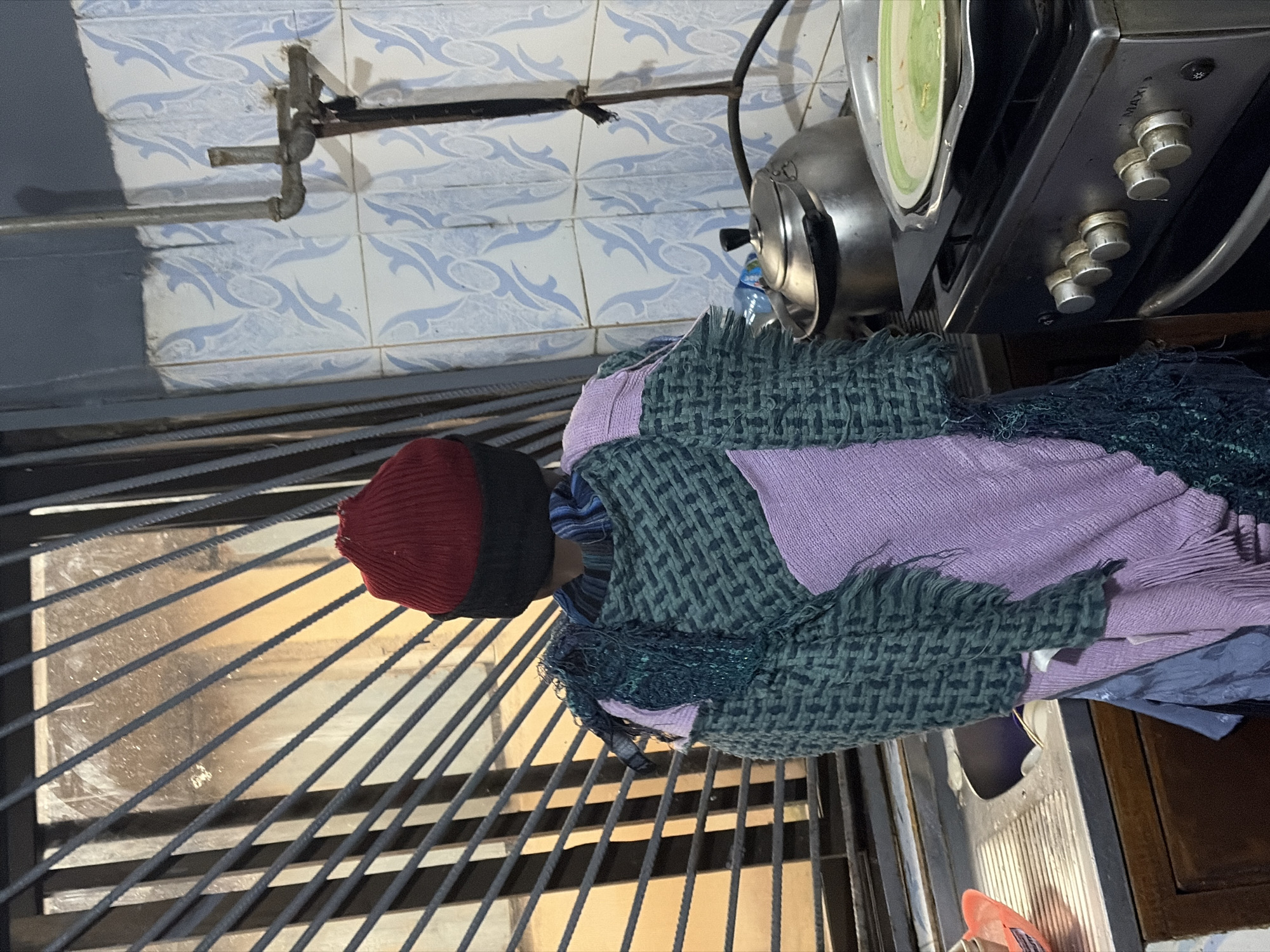 The image depicts a person wearing a red beanie and a multi-colored woven garment in what appears to be a kitchen setting. The background includes a metal-barred window with a view of an outdoor area, blue and white patterned tiles on the wall, a gas stove with a kettle and a plate on top, and a sink with a wooden cabinet below. The person is facing away from the camera, suggesting they are standing or engaged in an activity within the kitchen. The overall impression is that of an indoor domestic scene, possibly in a residential area of Jos, Nigeria.