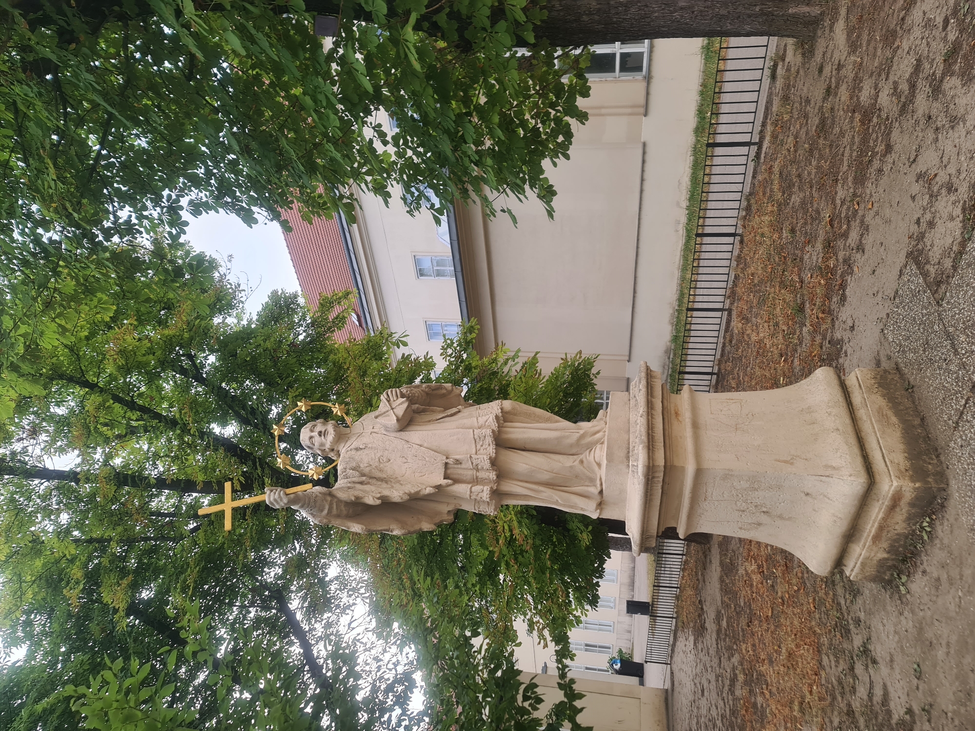 The image shows a statue of a male figure holding a golden cross above his head and a book in his other hand. The statue is made of light-colored stone and stands on a pedestal. It is surrounded by trees with green leaves, and a light-colored building with a red tiled roof is visible in the background. The ground is covered with dirt, dry leaves, and some grass. A black metal fence runs horizontally across the middle ground of the image. The statue is located in front of a building in Vienna, Austria, at the St. Mary of the Angels Church (Kirche Maria vom Siege).