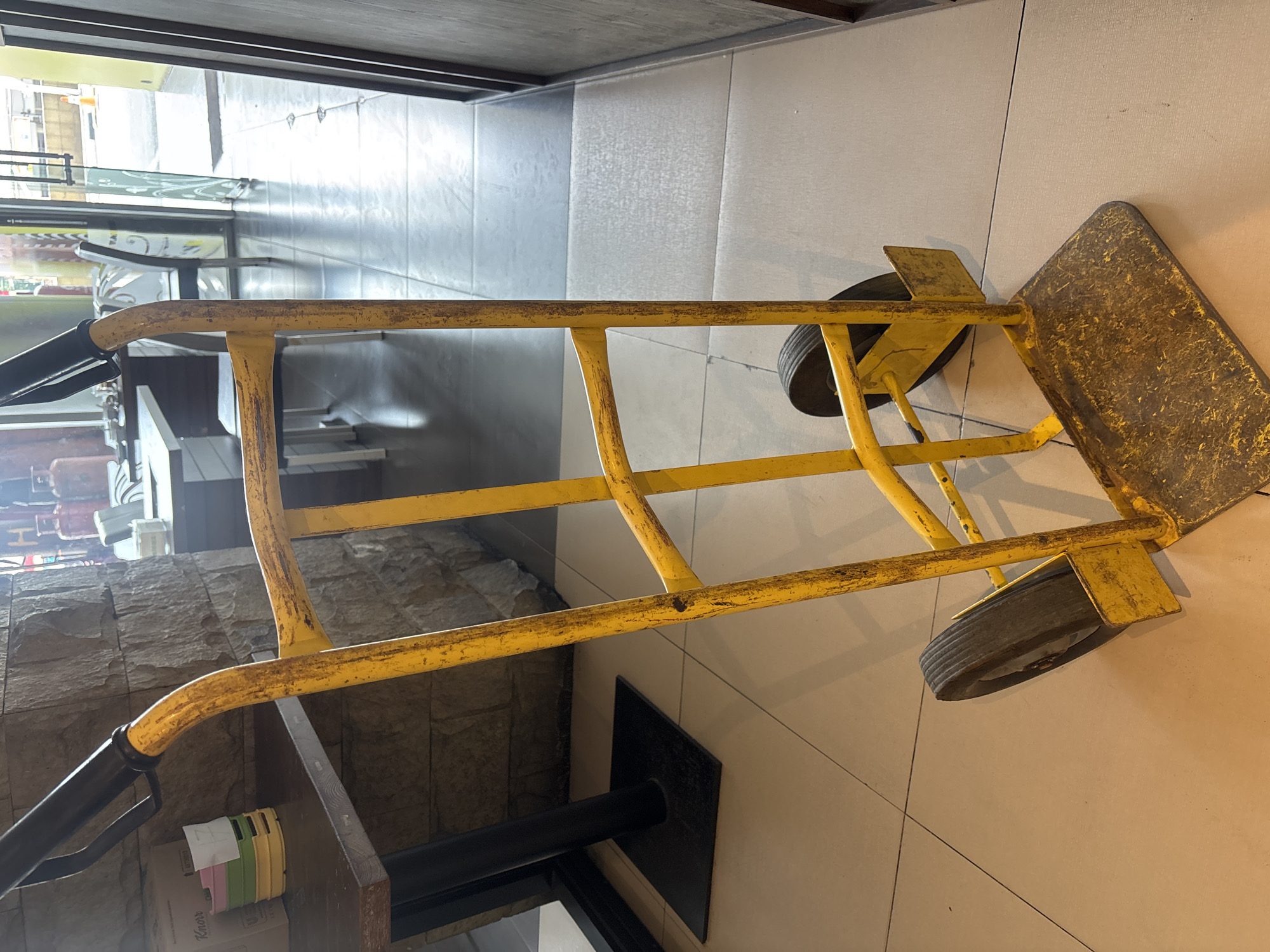 A yellow hand truck is positioned vertically on a tiled floor. The hand truck has two black rubber wheels and a metal platform at the base, with two black handles at the top. The hand truck appears worn and slightly rusty in places. Behind the hand truck, a stone wall is visible, and further back, a glass door leads to an outdoor area where a traffic cone can be seen. The scene suggests a commercial or restaurant setting, likely within Pekanbaru, Indonesia, given the context. The lighting is moderate, casting some shadows on the floor. There are no people or animals present in the image.