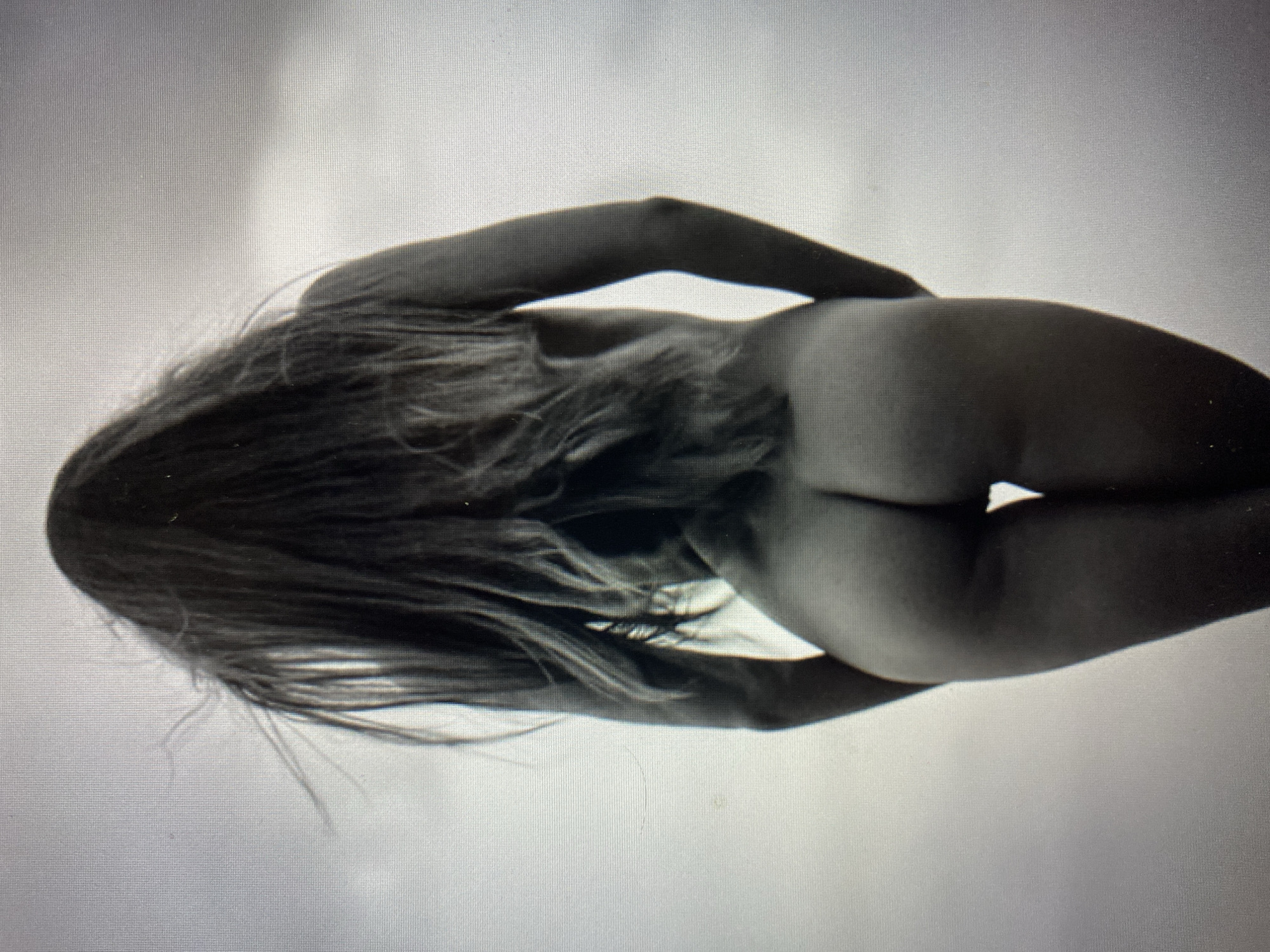 The image shows a person from behind, with long hair flowing. The person is facing away from the camera, and their back and buttocks are visible. The background is a blurred, light-colored sky or wall. The image is in black and white and has a grainy texture. There are no discernible features of a specific location in the city of Unknown, Unknown.